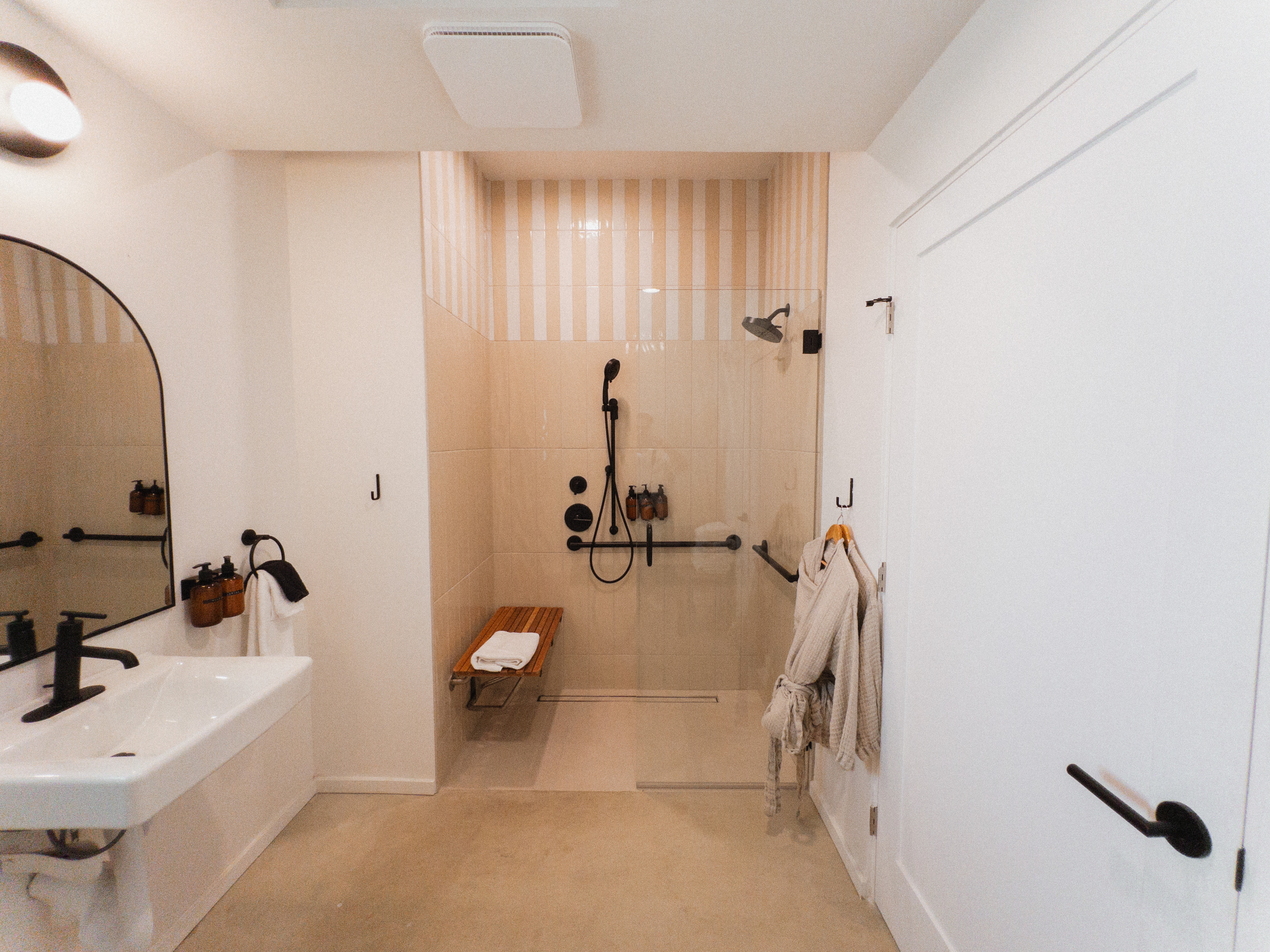An accessible bathroom featuring a vanity with an arched mirror and a spacious, glass-enclosed roll-in shower with a shower bench, grab bars, and light-colored vertical striped accent tile.