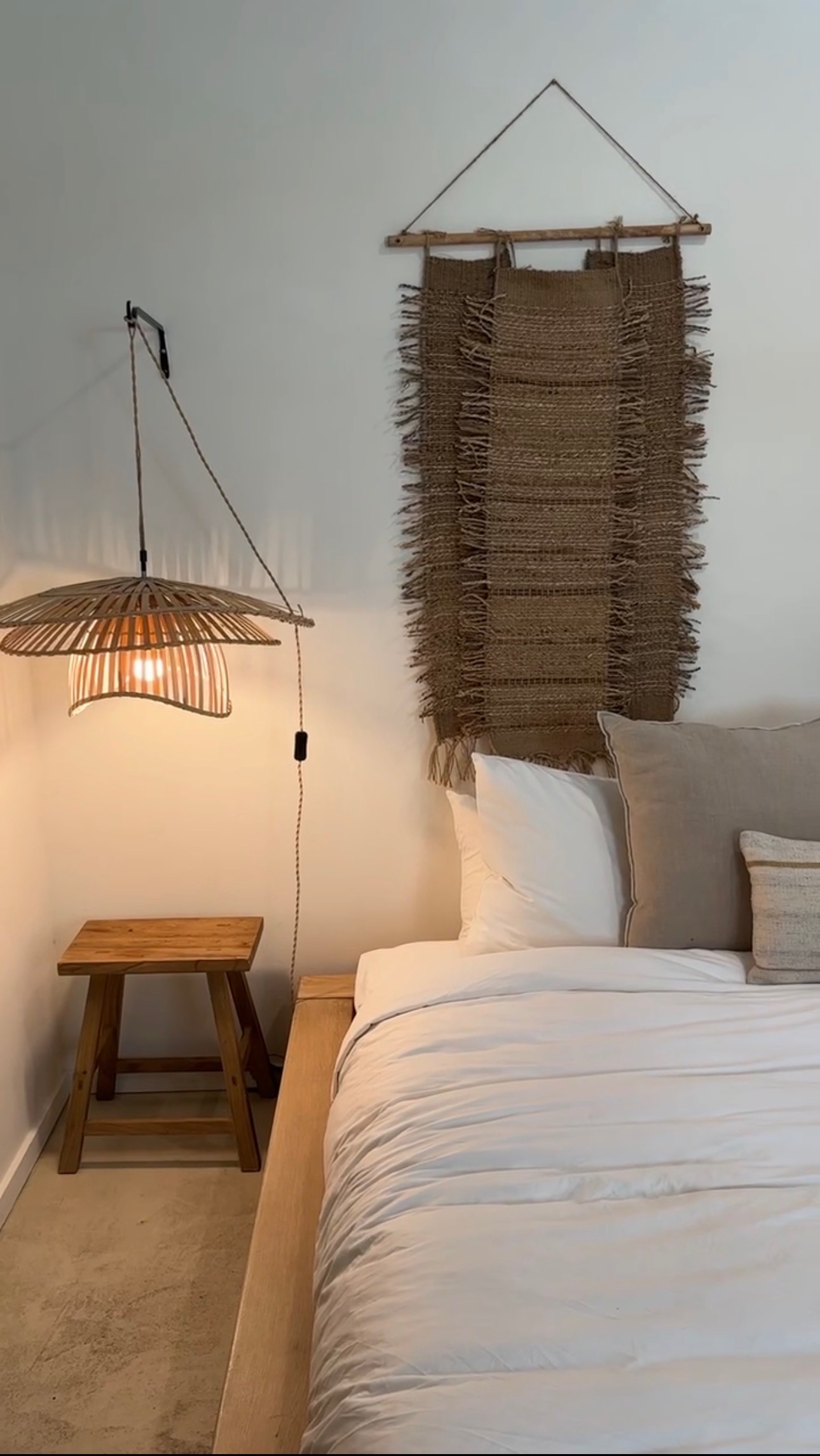 A vertical shot of a minimalist bedroom featuring a light wood platform bed with white linens. The wall is decorated with woven art and a hanging raffia pendant lamp over a rustic wood side table.