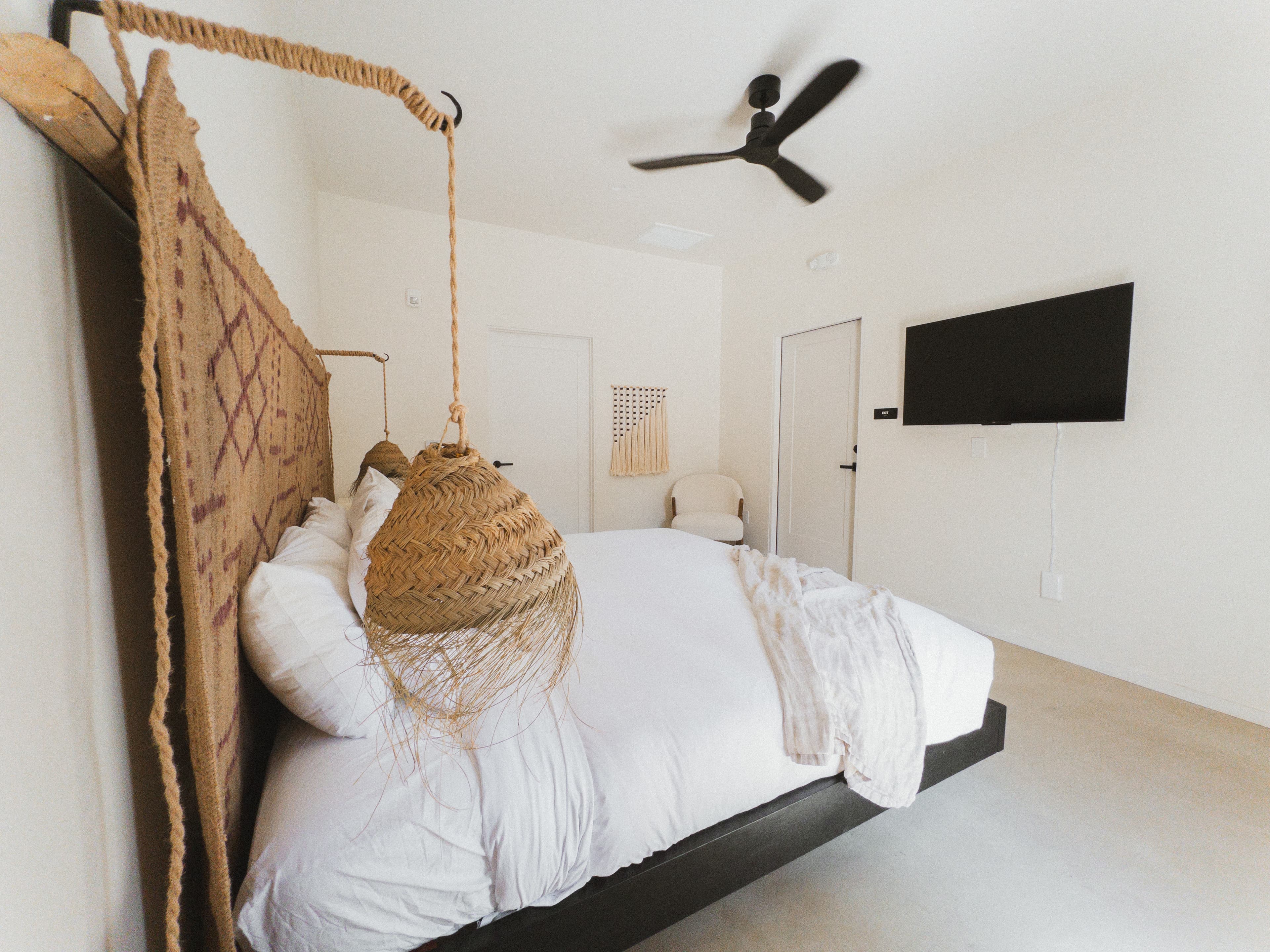 A hotel bedroom featuring a platform king bed with a tall, woven headboard. The room includes a wall-mounted TV, a black ceiling fan, and a hanging raffia light fixture.