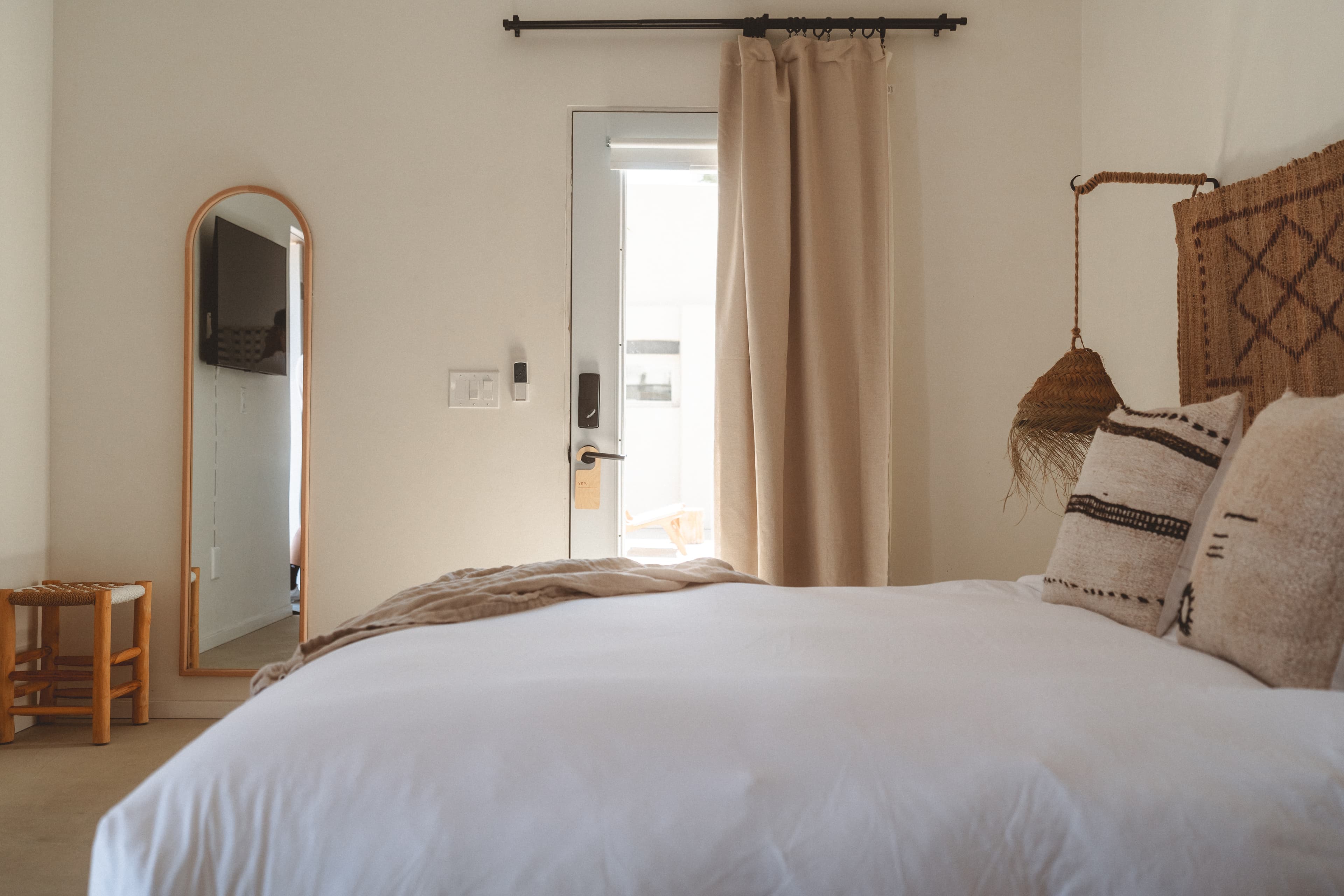 A bright hotel room featuring a king bed with layered linens, a woven headboard, and a full-length arched mirror reflecting a wall-mounted TV. A door with sheer curtains leads outside.