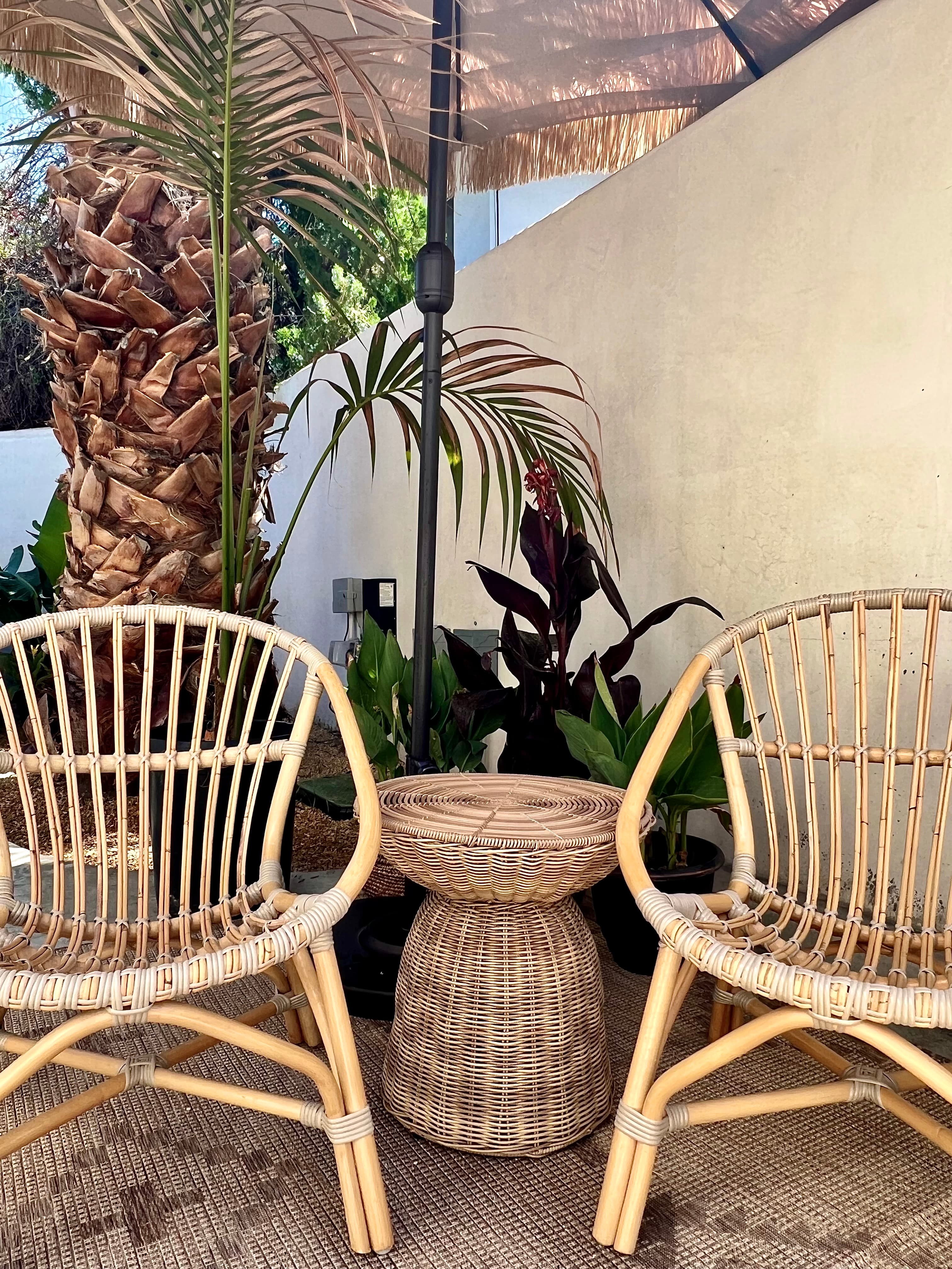 Two light-toned rattan armchairs and a woven side table sit on a patio. The area is lushly decorated with palm trees and tropical plants, shaded by a fringed umbrella.