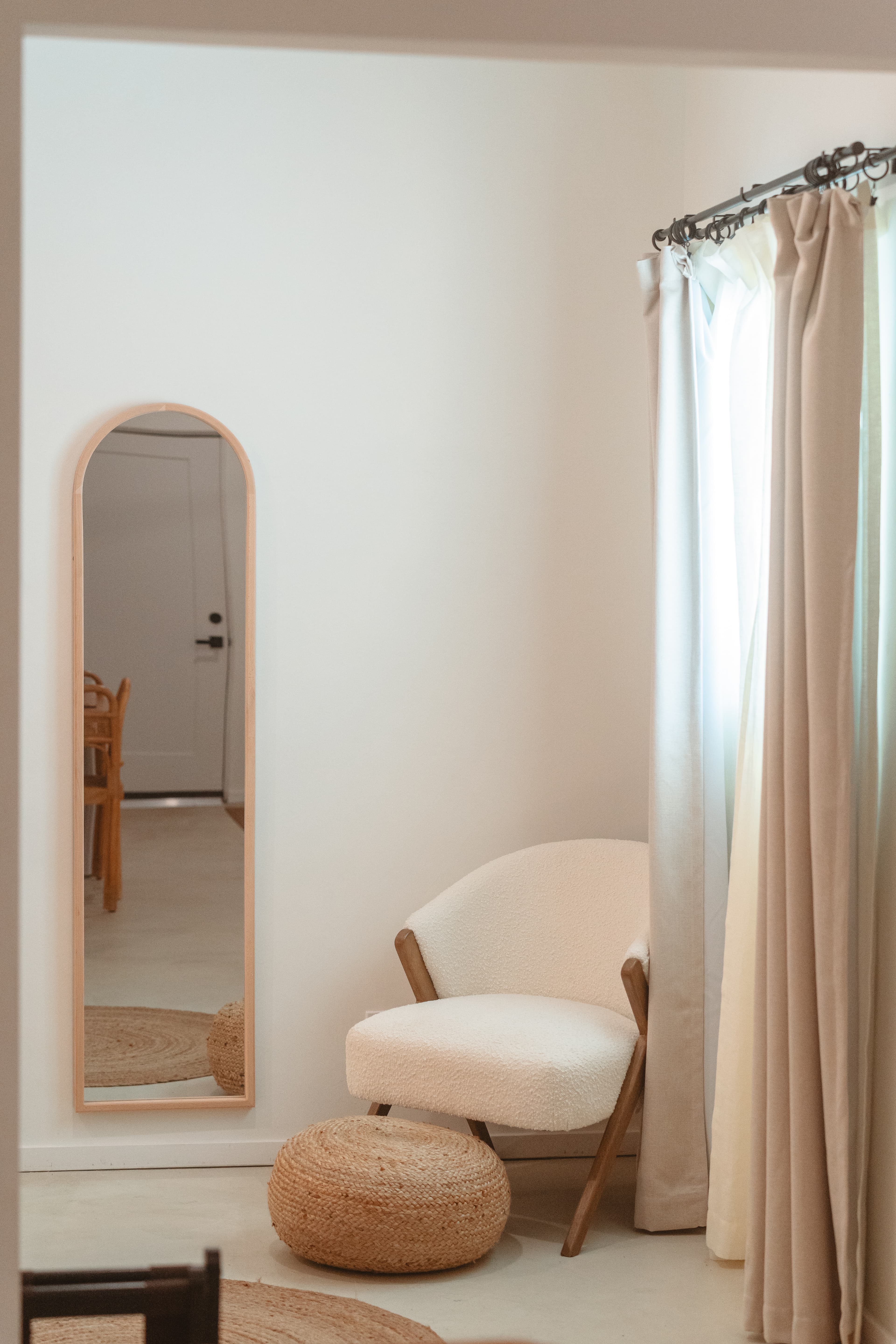 A bright room corner featuring a neutral-toned, upholstered accent chair with wood framing, a woven round ottoman, a full-length arched mirror, and floor-to-ceiling curtains.