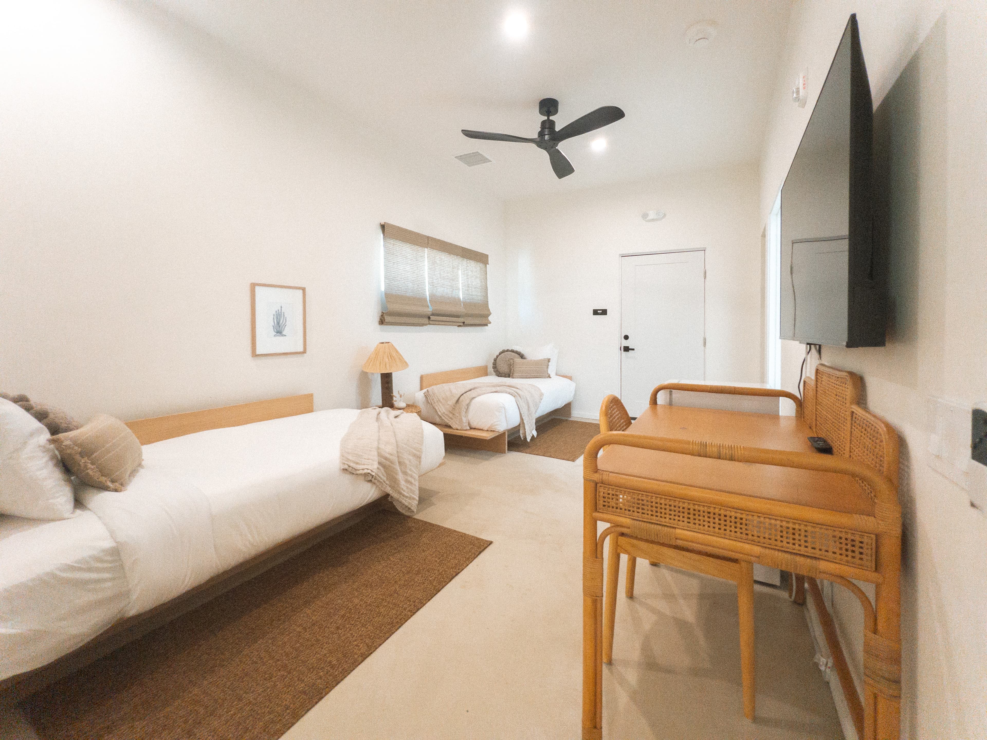 A bright hotel suite featuring two twin-sized daybeds, a rattan desk with a chair, a wall-mounted TV, and a black ceiling fan, all in a neutral, modern aesthetic.
