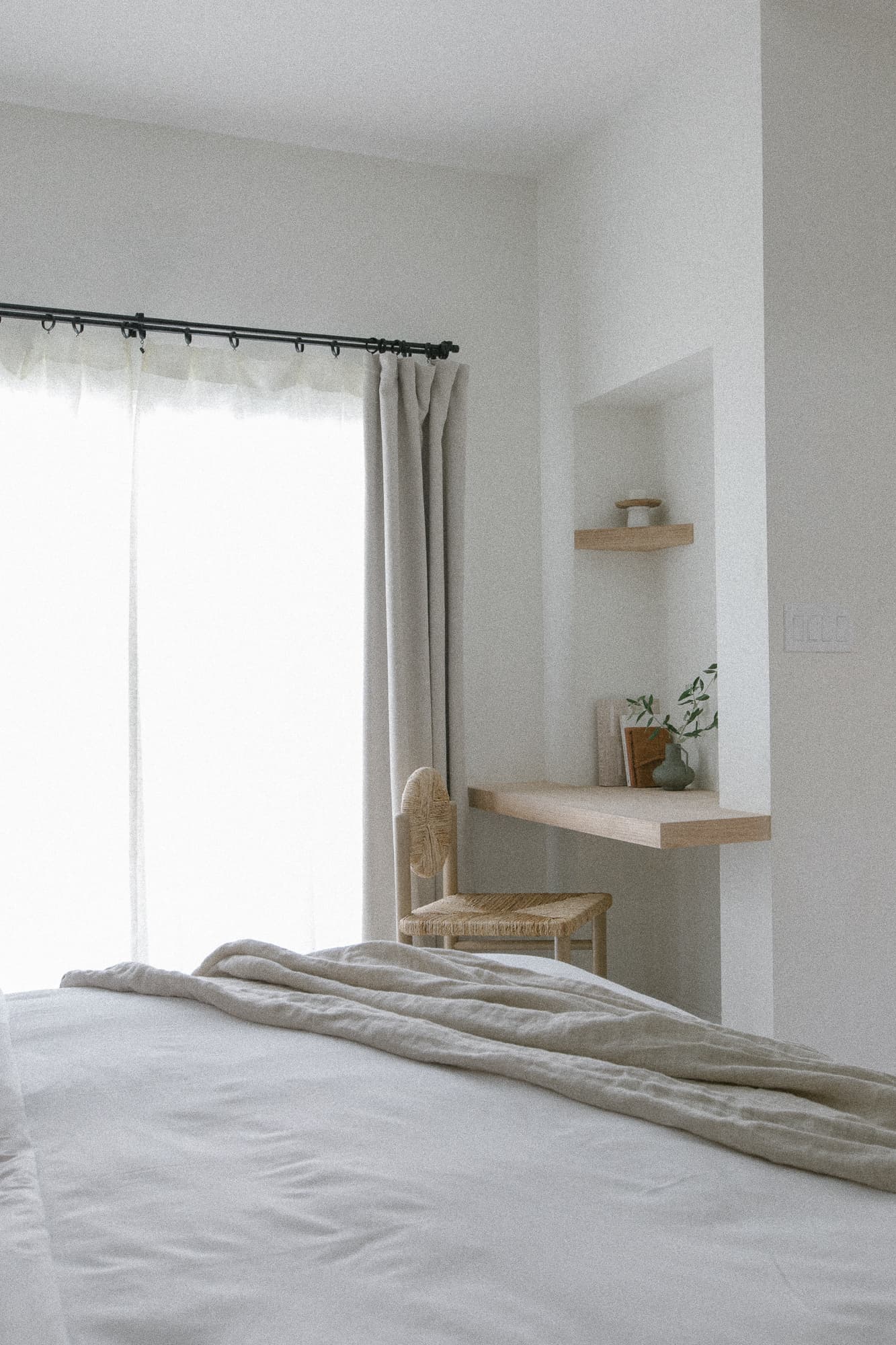 A bright bedroom with a bed covered in light linens, facing large sliding glass doors draped with neutral curtains. A built-in wooden desk/shelf area with a woven chair is visible in the corner.