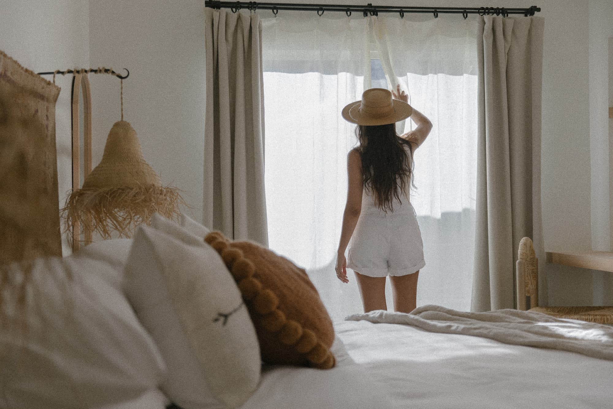 A woman with long dark hair wearing a straw hat and white romper stands at a bright window, drawing back a sheer curtain. The foreground shows a bed with textured, neutral-toned pillows.