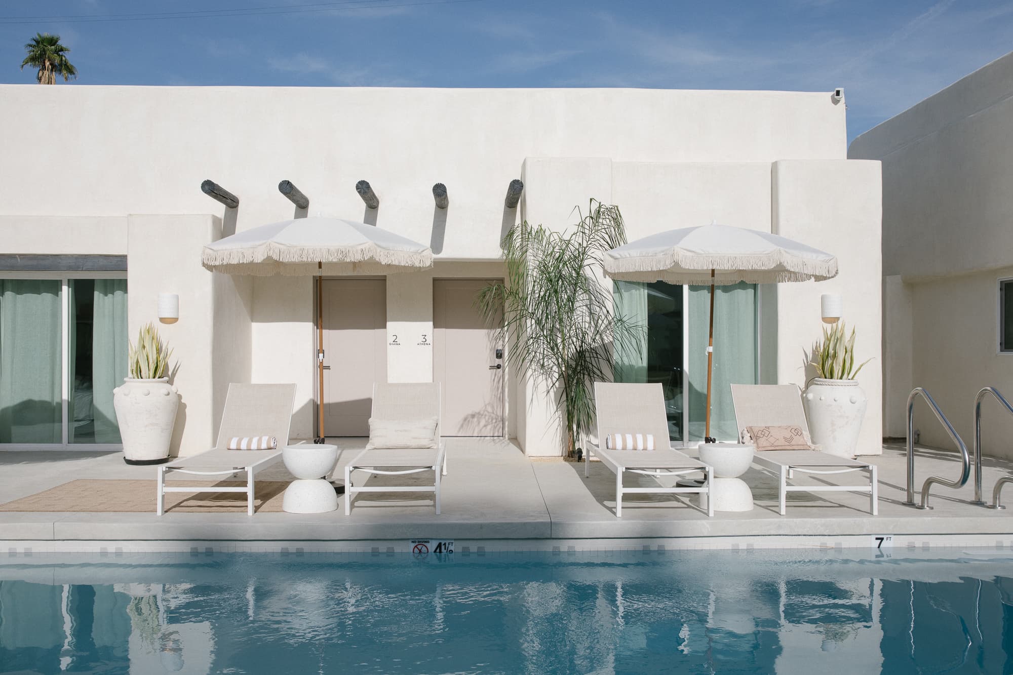 A bright, sunny view of a hotel pool with two poolside lounge chairs, each shaded by a white umbrella. The chairs face the white, minimalist exterior of the rooms.