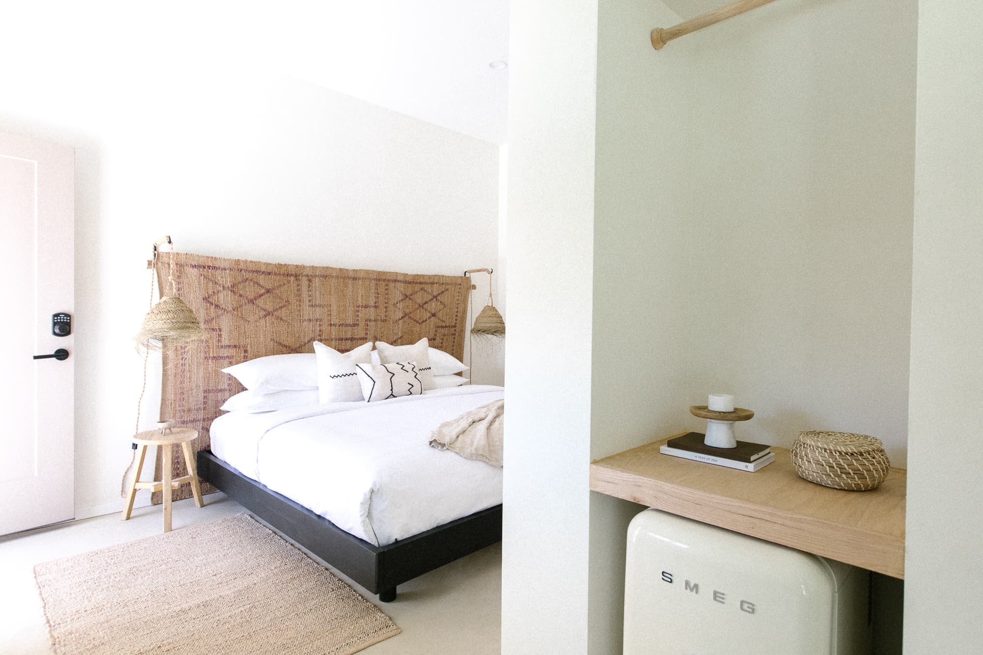A bright, minimalist hotel room featuring a king bed with a woven headboard, natural fiber rug, and a light-colored SMEG mini-fridge/mini-bar area.