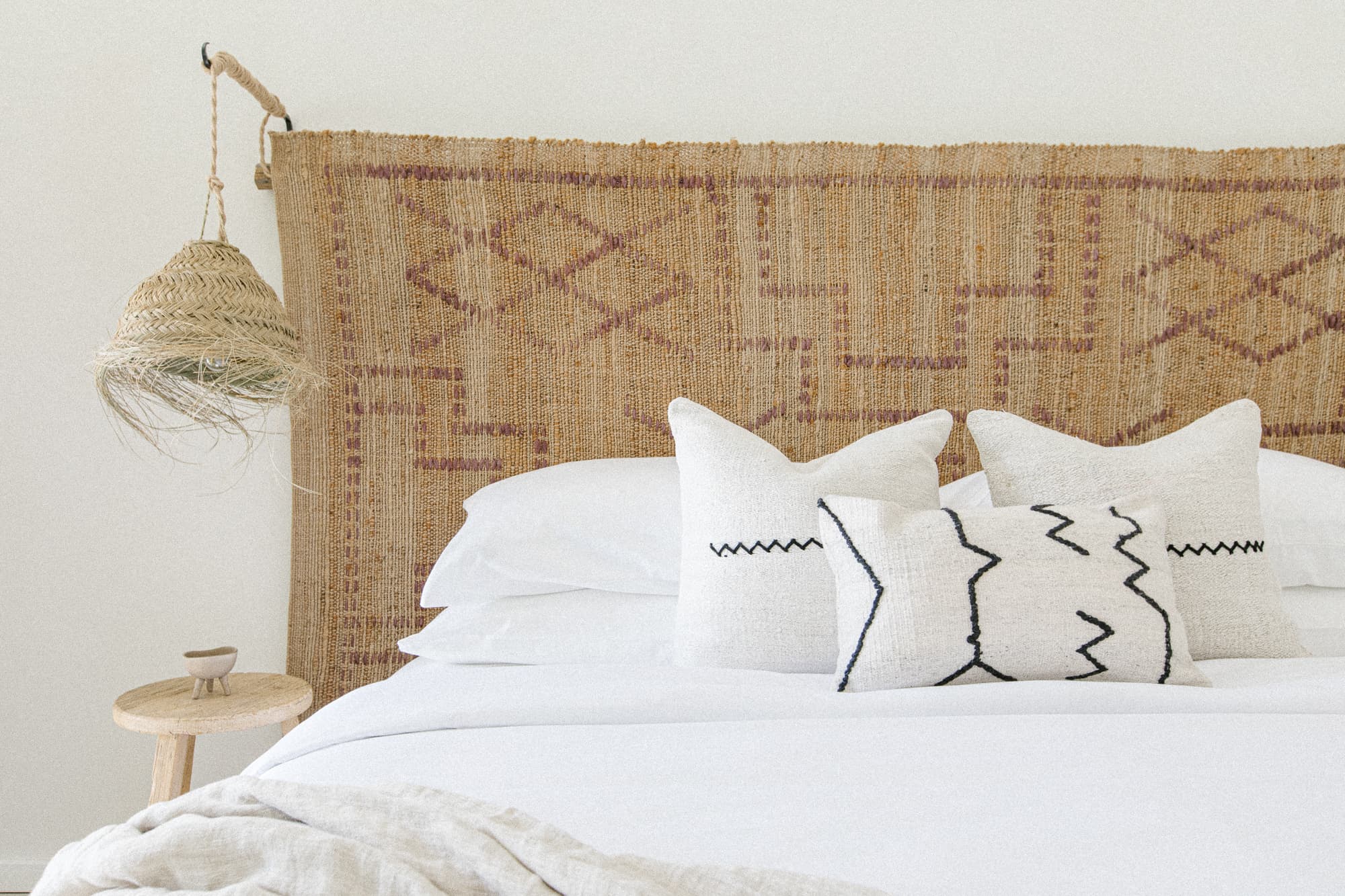 A close-up of a bed featuring a textured, woven headboard and crisp white linens. Accent pillows have geometric black and white stitching. A rattan pendant light hangs above a wooden side table.