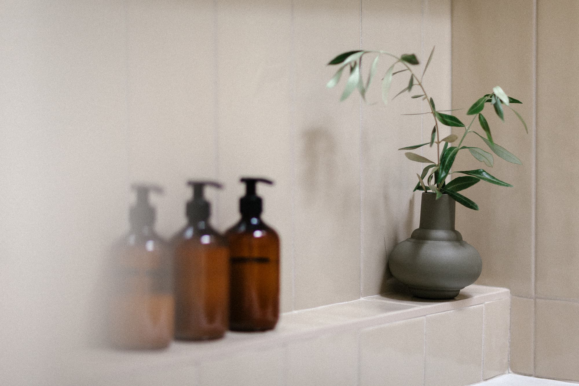 A close-up shot of a bathroom niche with tan tiles. It holds three amber pump bottles and a small, matte gray-green vase with fresh green foliage.