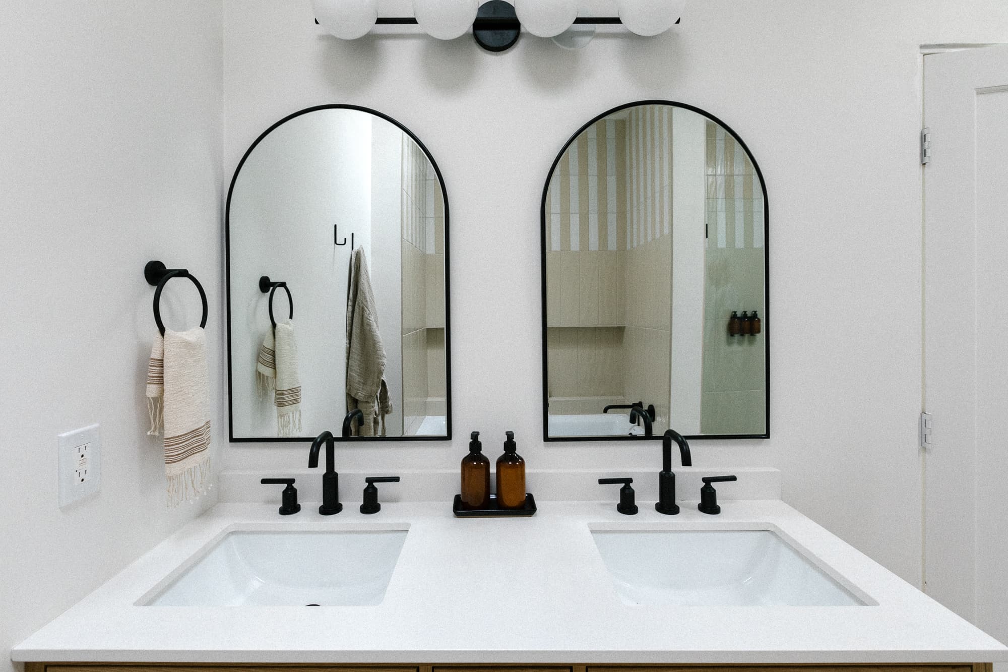 A modern double vanity with arched mirrors framed in black. The counter features black faucets, a white countertop, and dark amber amenity bottles.