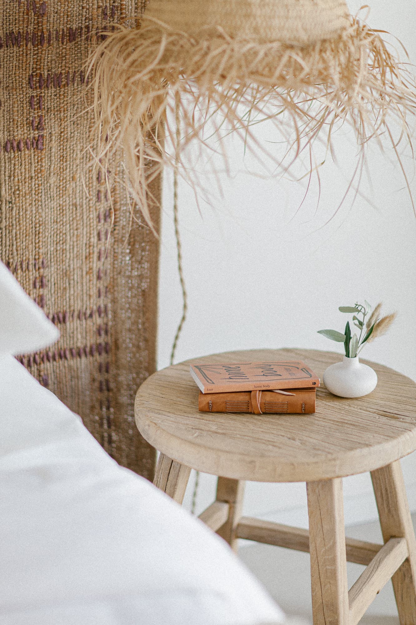 A close-up of a rustic wooden side table next to a bed with white linens. The table holds a stack of books and a small white vase with greenery, all under a woven raffia light fixture.