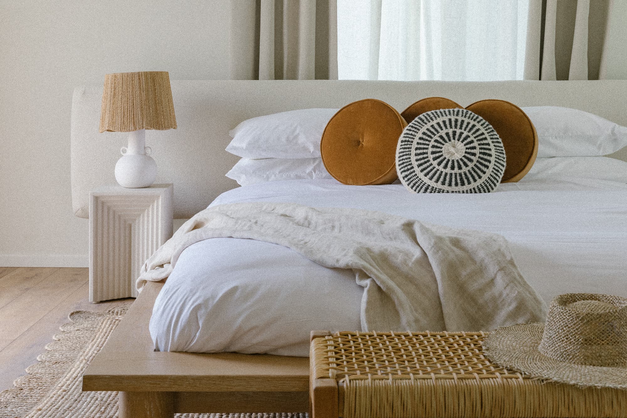 A close-up of a modern bed with a cream upholstered headboard and white linens. It features mustard and embroidered round accent pillows, a woven bench, and a lamp with a rattan shade.