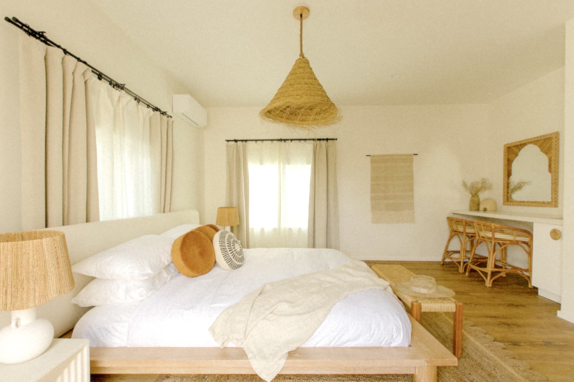 A bright hotel room with a wooden platform bed, white linens, and neutral curtains. It features a woven light fixture, a rattan desk with a carved mirror, and a calming, natural aesthetic.