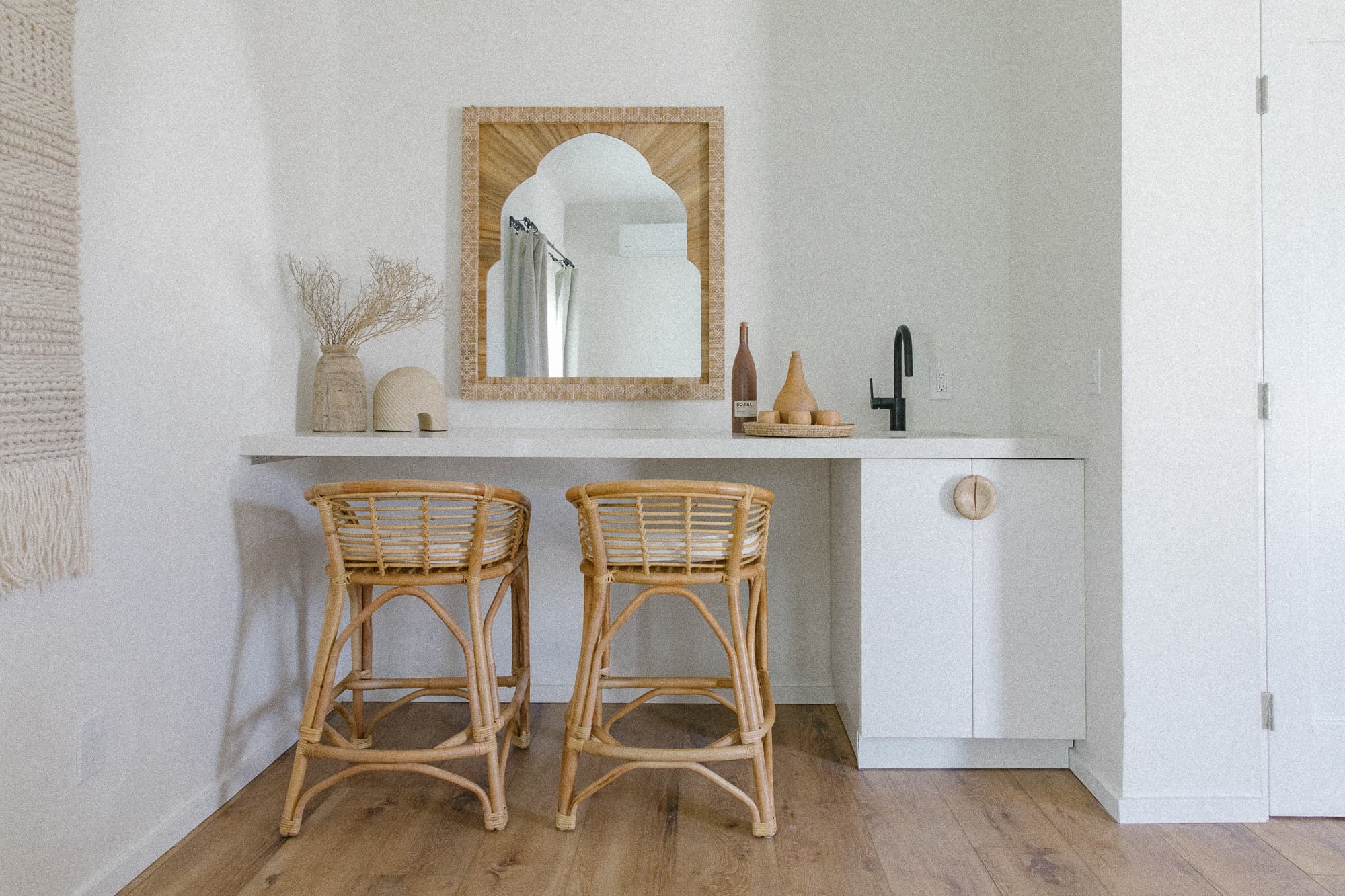A bright, minimalist wet bar area featuring a white countertop, black sink and faucet, and two rattan bar stools. An arched wooden mirror hangs above the counter.