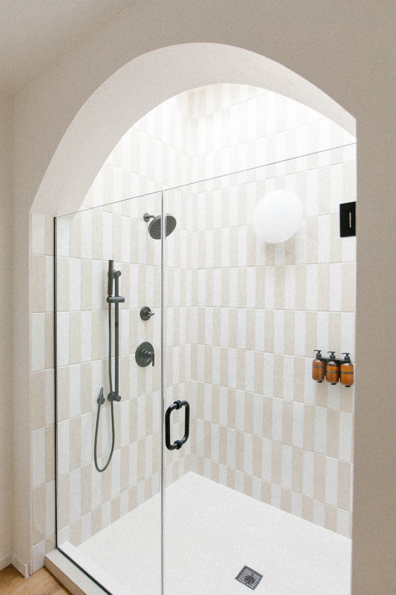 A walk-in shower with a glass door set under an arched opening. The interior features beige and white vertical striped tile, black fixtures, and a white globe light fixture.