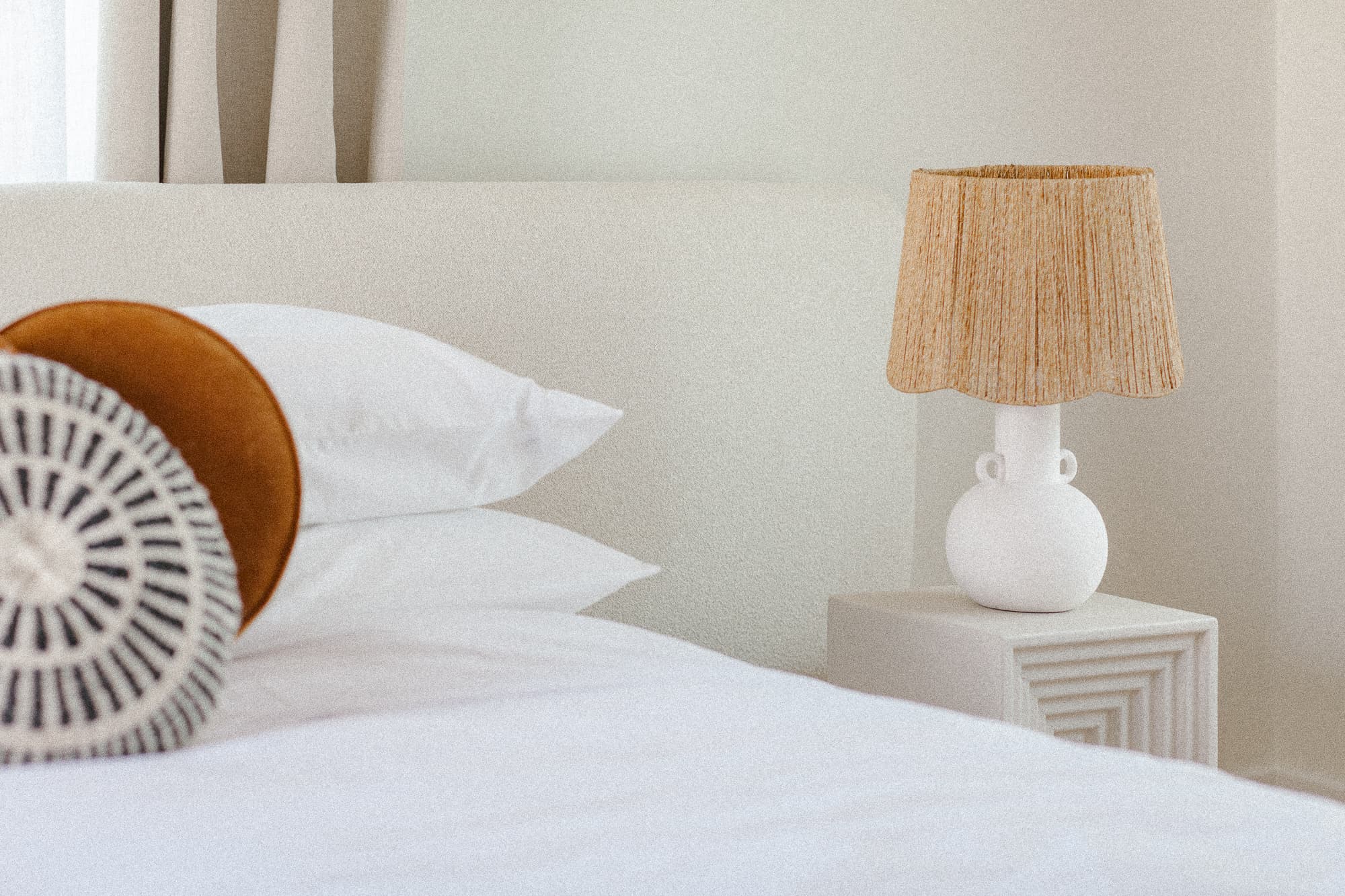 A close-up of a bed with white linens and decorative pillows. A white ceramic table lamp with a natural, textured lampshade sits on a minimalist white nightstand.