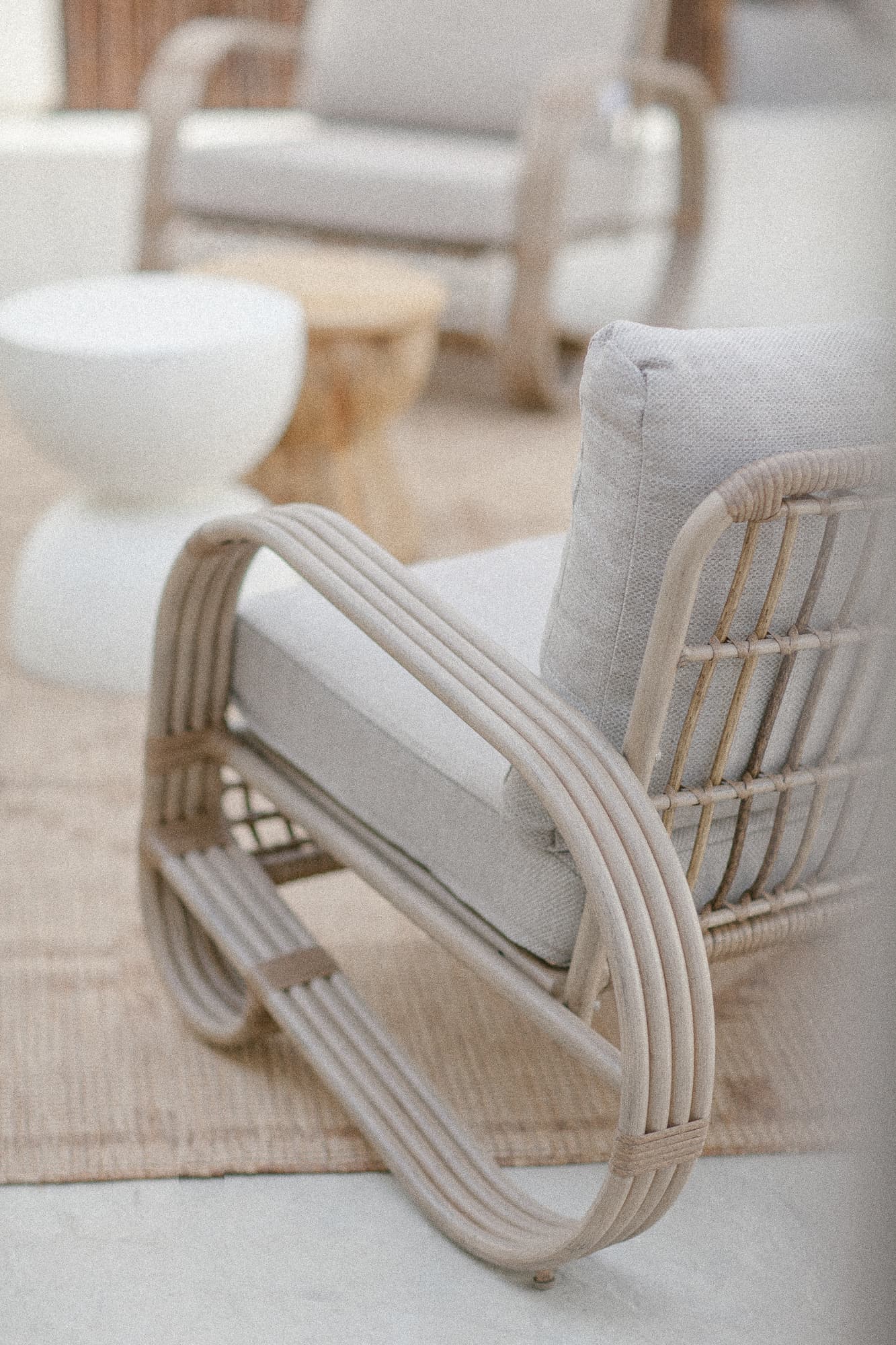 A close-up shot of a contemporary outdoor armchair with a light-toned curved rattan frame and neutral cushions, set on a beige patterned rug next to sculptural side tables.