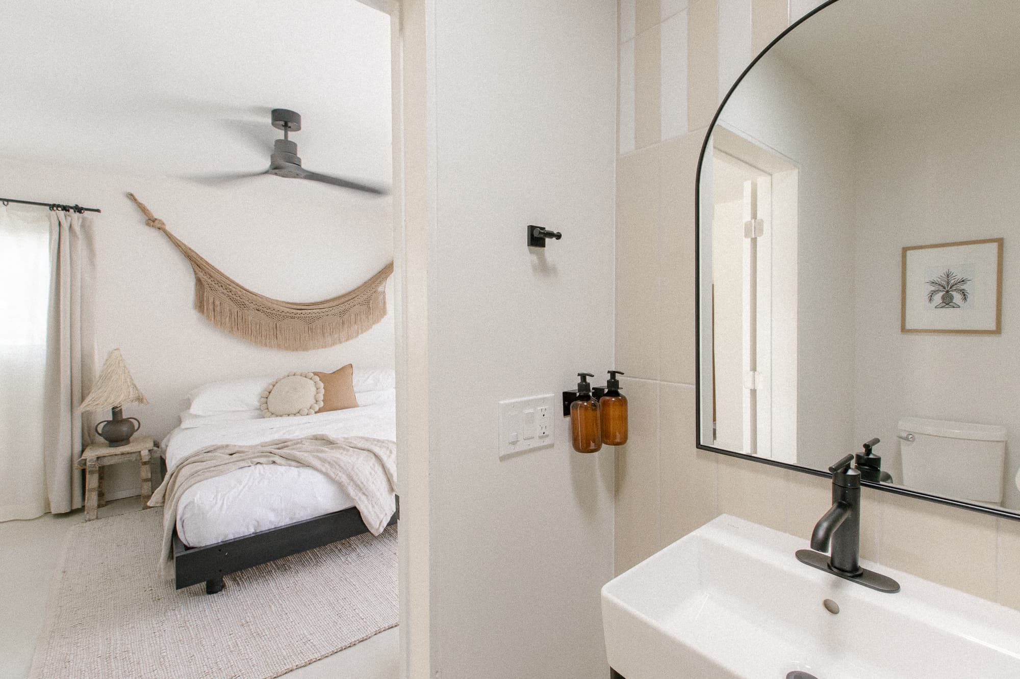 The view from a bright bathroom, featuring a vanity with an arched mirror and a striped accent wall, into the adjacent bedroom with a large bed and a macrame wall hanging.