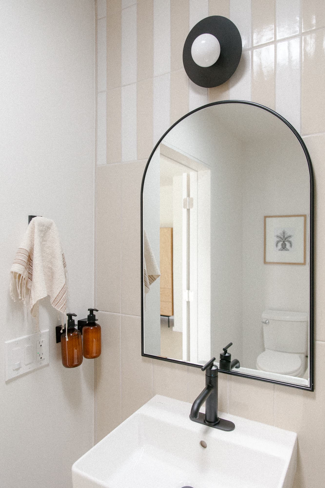 A stylish bathroom featuring a white sink, a black arched mirror, and a wall of beige and white striped tile. Black fixtures and amber soap dispensers complete the look.