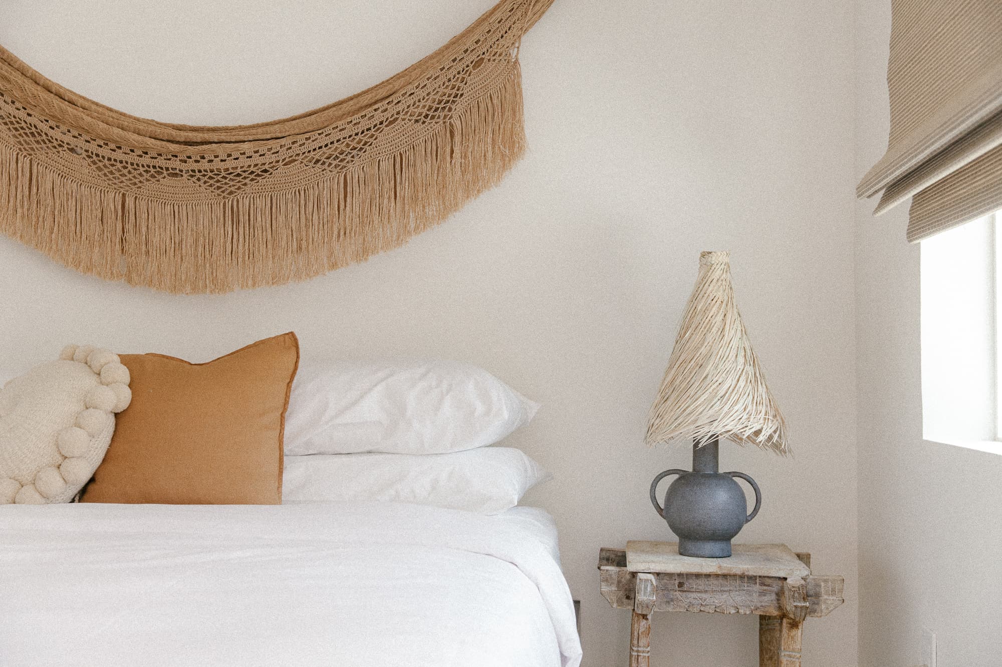 A close-up of a bed with white linens and warm accent pillows. Above the bed is a woven macrame wall hanging, and a unique lamp with a raffia shade sits on a rustic wooden side table.