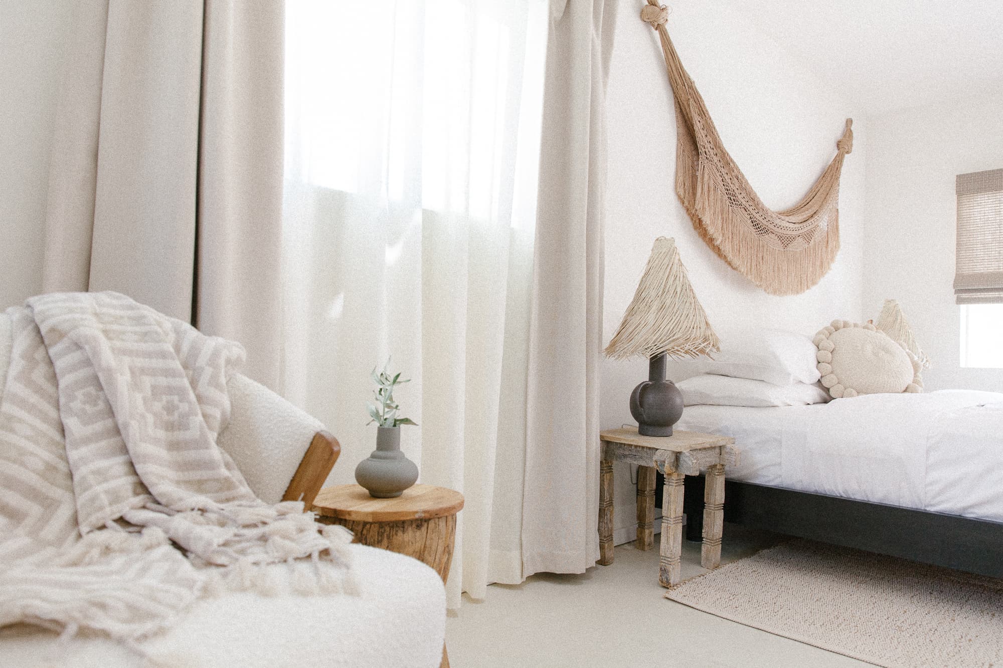 A bright, tranquil bedroom corner featuring an upholstered accent chair with a patterned throw, natural wood side tables, and large windows with sheer curtains.
