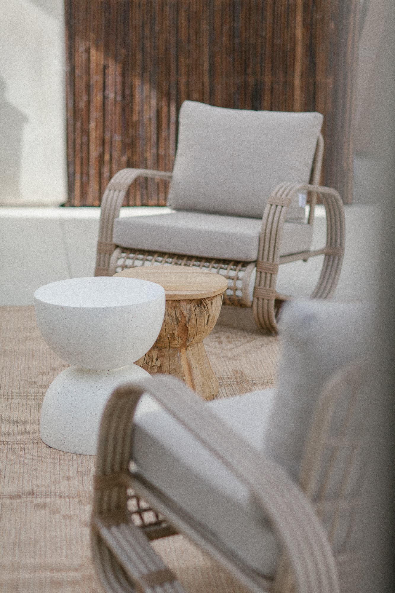 A close-up of an outdoor seating area featuring two modern, curved rattan chairs with neutral cushions and two unique side tables-one white stone, one natural wood.