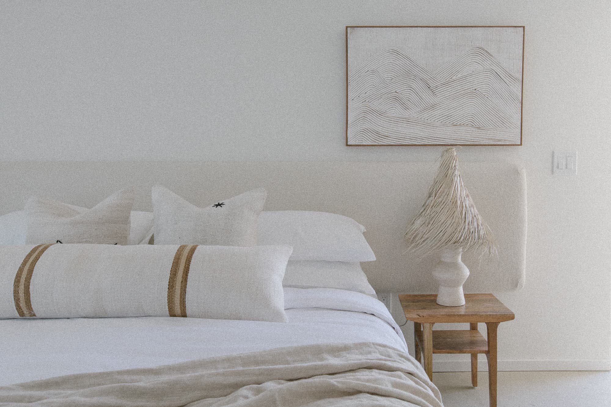 A bright, minimalist bedroom featuring a bed with a cream upholstered headboard, white linens, and neutral accent pillows. A textured lamp sits on a wooden side table beneath abstract wall art.