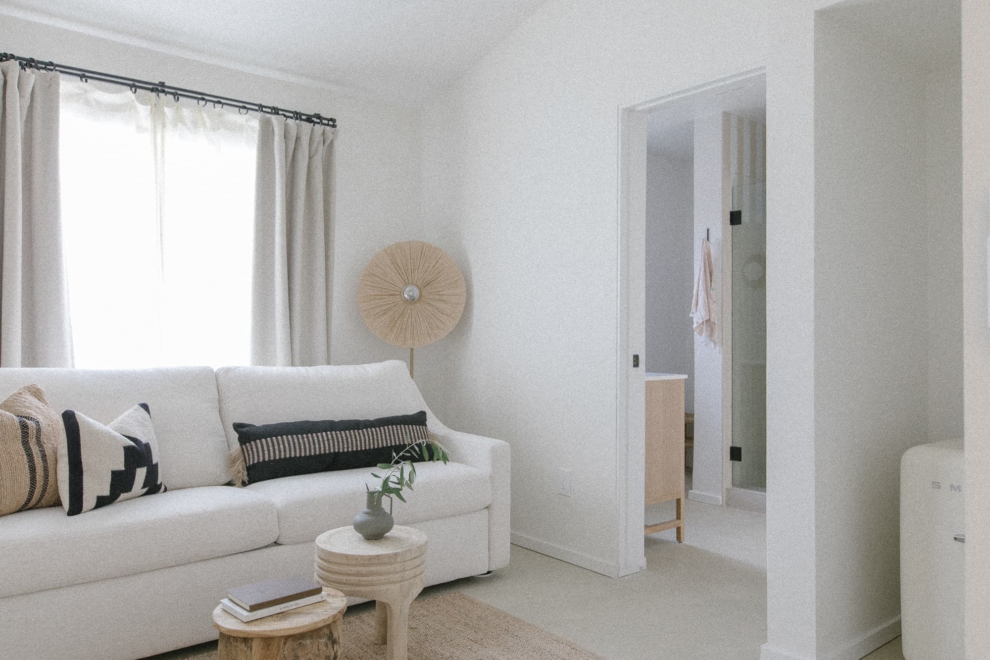 A bright, modern living area with a white sofa, neutral accent pillows, and natural wood coffee tables. An open doorway leads into the light-filled bathroom.
