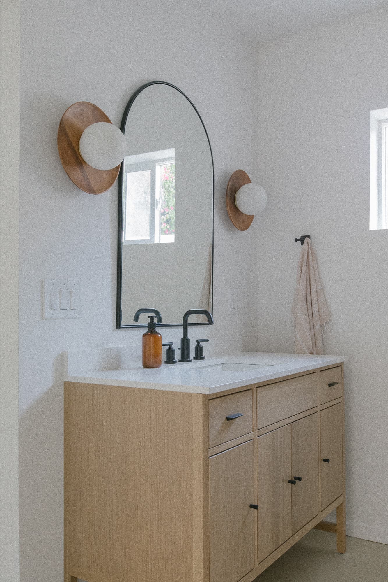 A modern bathroom vanity with a light wood cabinet, white countertop, and black fixtures. An arched mirror is flanked by mid-century modern style sconces with wood backs.