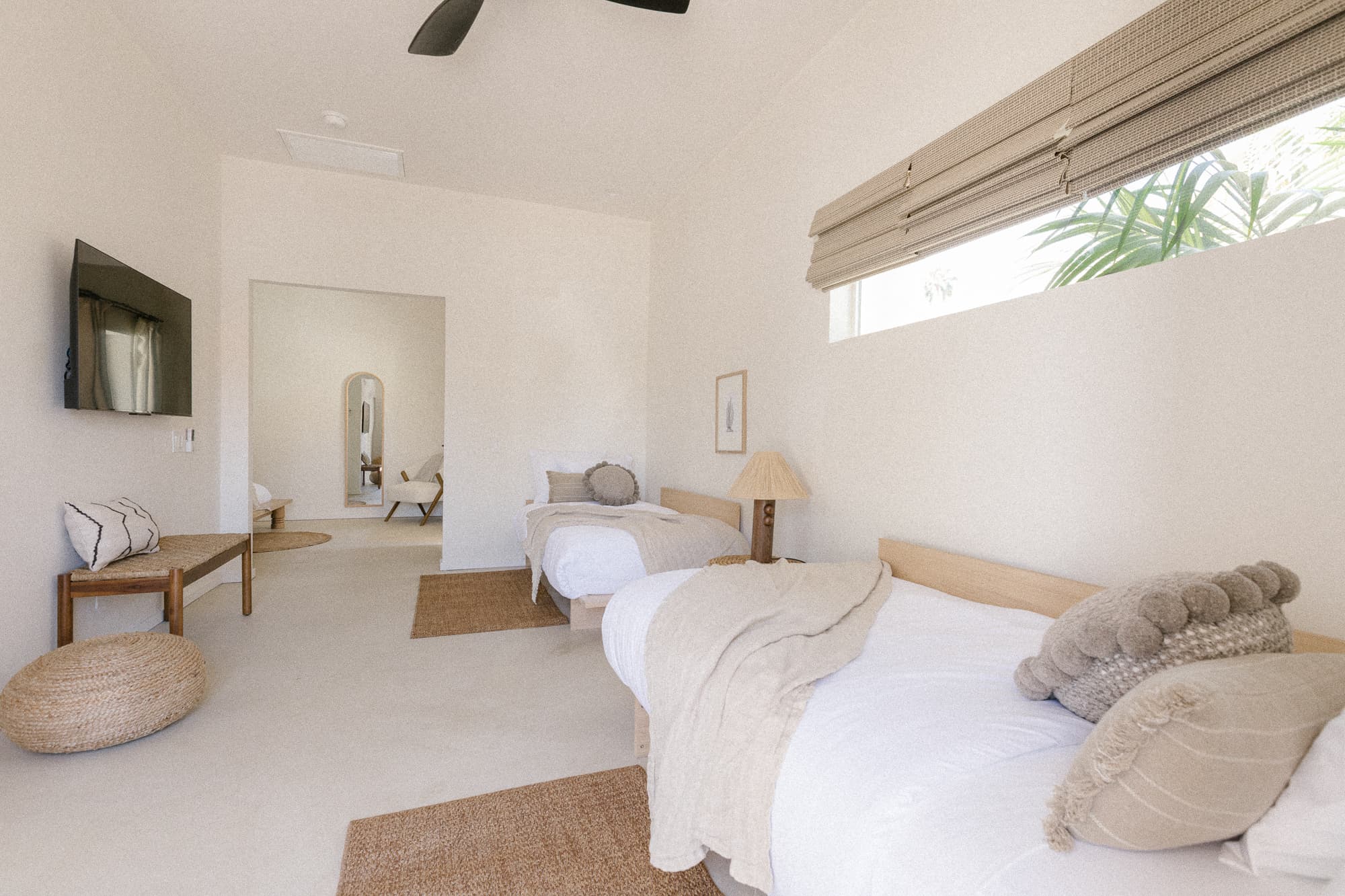 A bright, spacious suite featuring two wooden beds with neutral linens, a wall-mounted TV, and a long horizontal window. An adjoining room is visible through an archway.