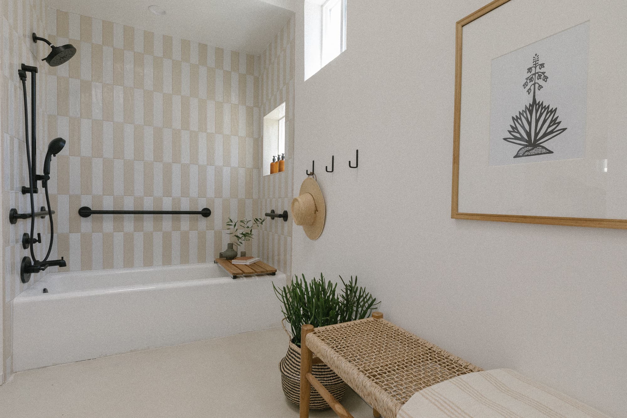 A bright, spa-like bathroom featuring a soaking tub with beige and white striped vertical accent tiles, black fixtures, and a woven bench with planters.
