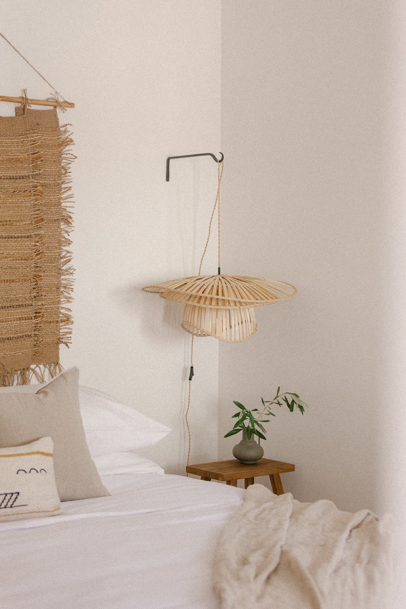 A close-up of a bed corner featuring a light woven wall hanging and a raffia pendant light suspended over a small wooden side table with a vase. The bed has layered white and neutral linens.