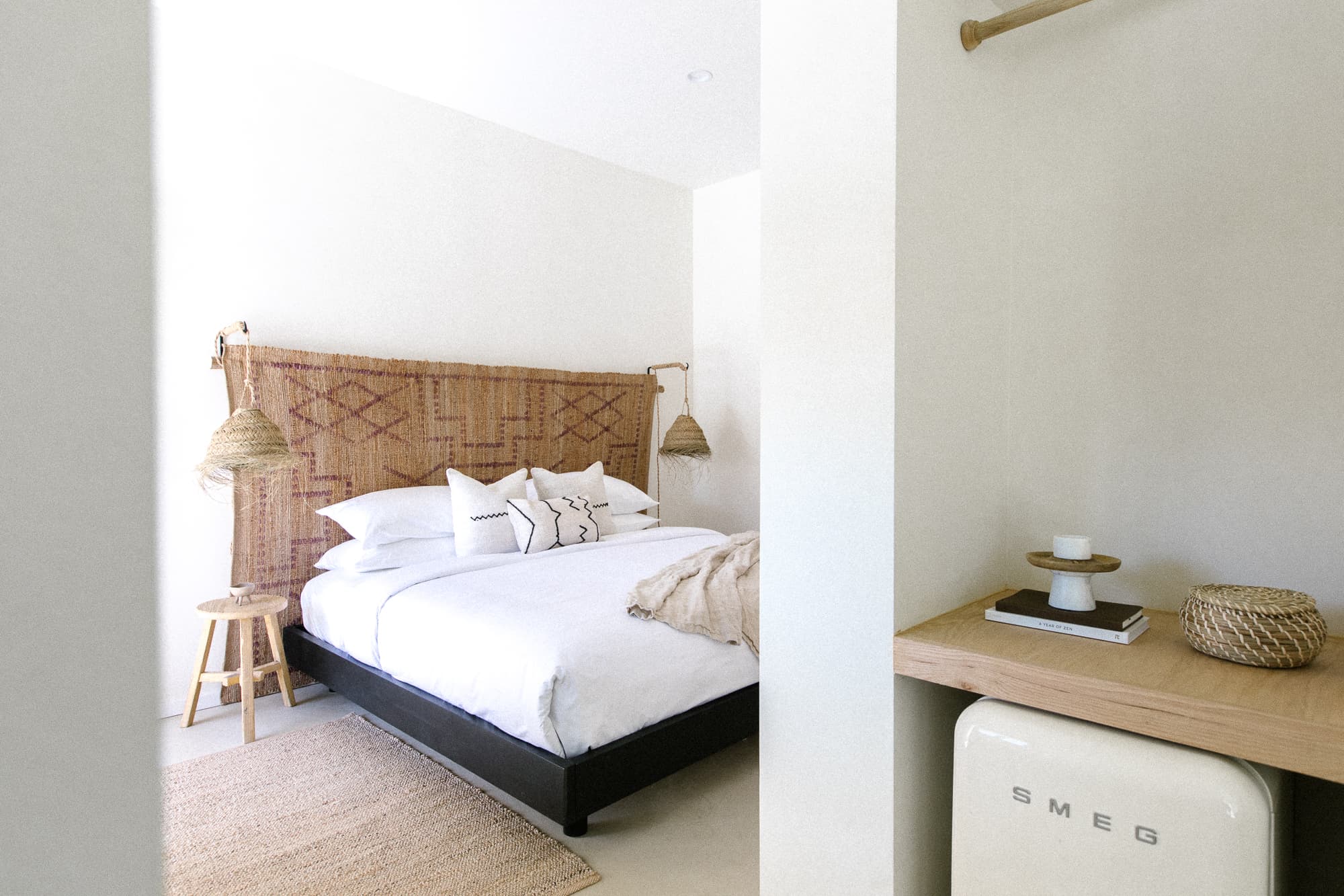 A view into a bright hotel room featuring a king bed with a woven headboard and neutral textiles. A light-colored SMEG mini-fridge/mini-bar area is visible in the foreground alcove.