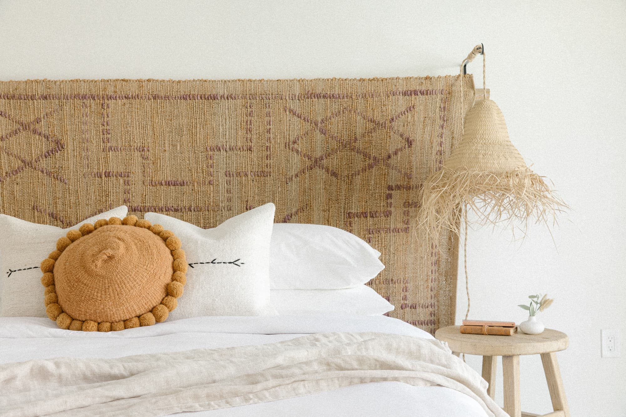 A close-up of a bed with a large woven textile headboard and white bedding. Accent pillows include a round rust-colored one and white ones with embroidery. A raffia pendant light hangs above a wooden side table.