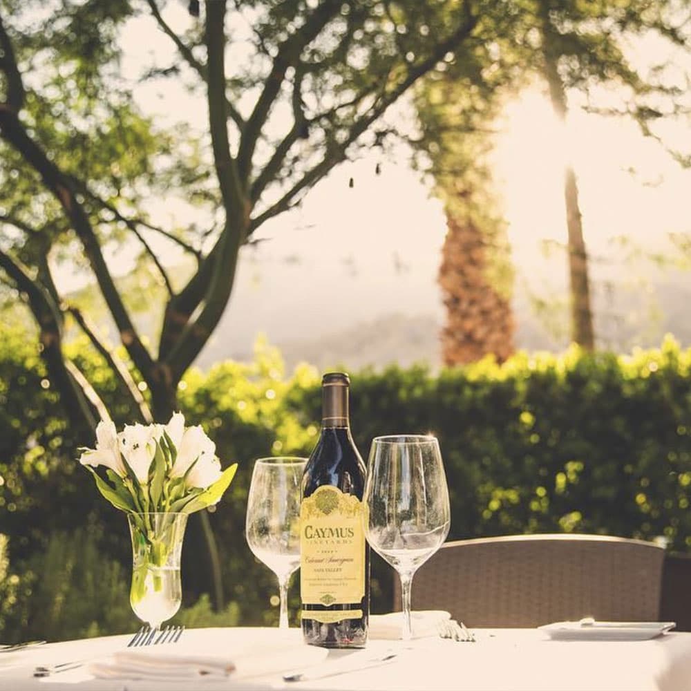 A bottle of Caymus wine sits on a table set with glasses and a vase of flowers, surrounded by greenery in soft sunlight.