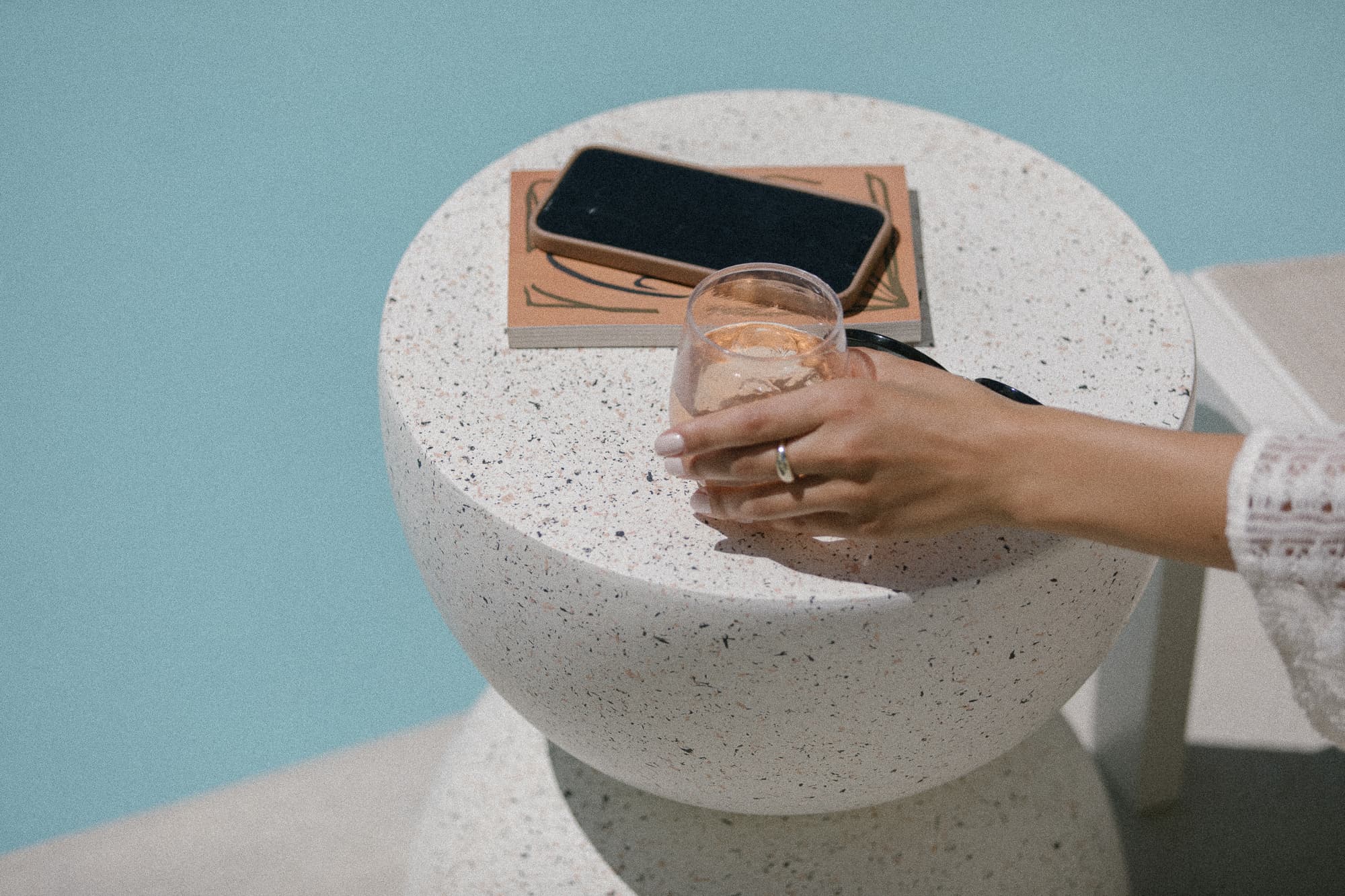 A hand holding a glass rests on a textured white table beside a phone and a notepad, with a pool in the background.