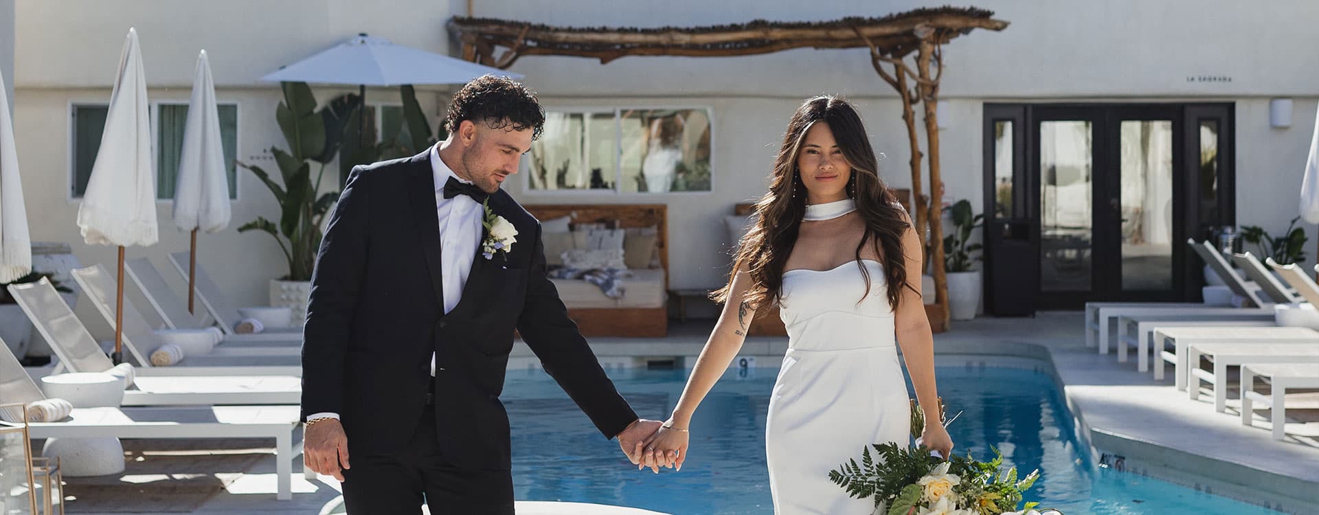 A couple in formal attire holds hands while walking by a pool at a sunny venue.