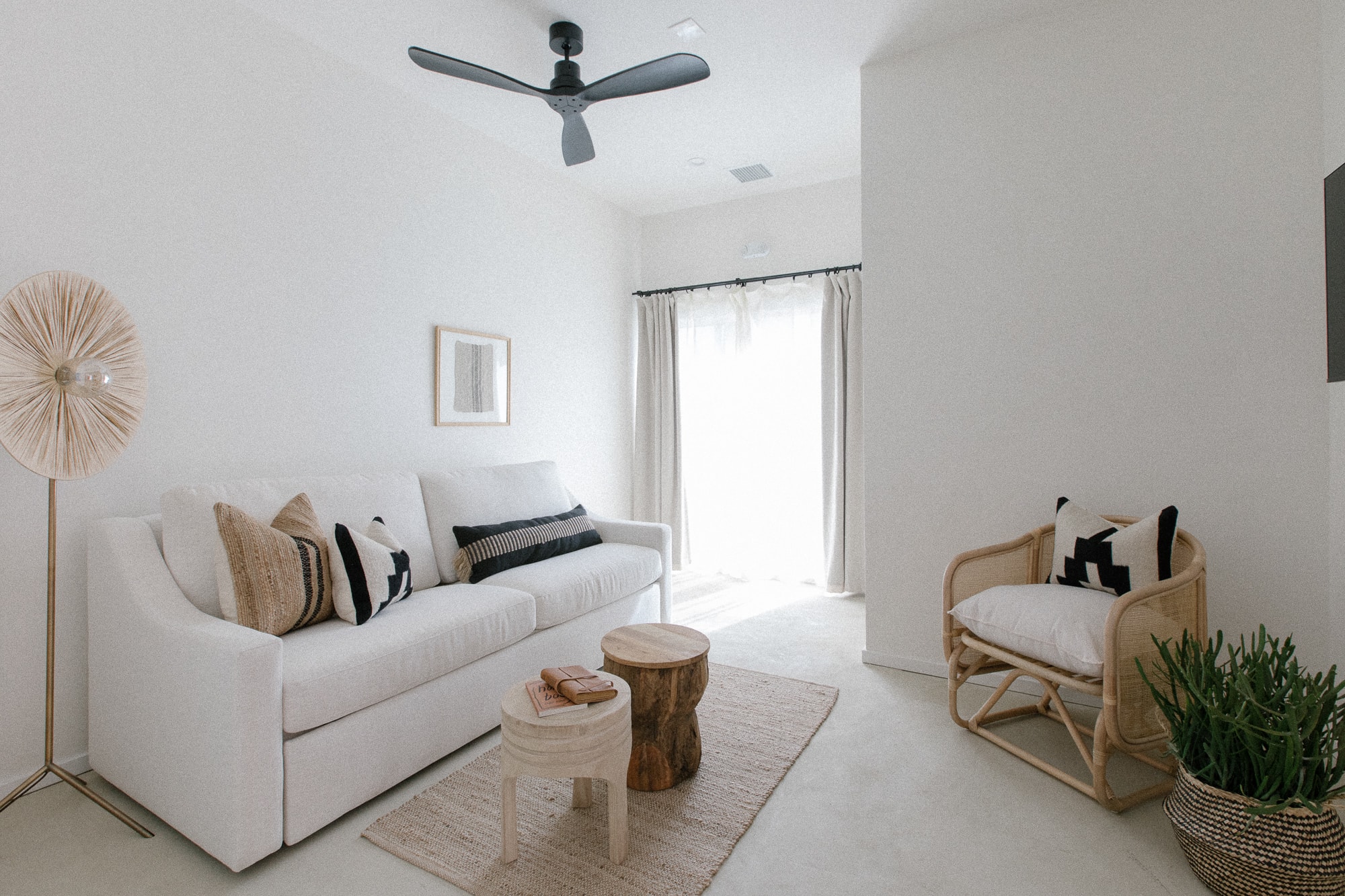 A bright, minimalist living space, complete with a white sofa, woven accent chair, natural wood tables, and direct outdoor access.