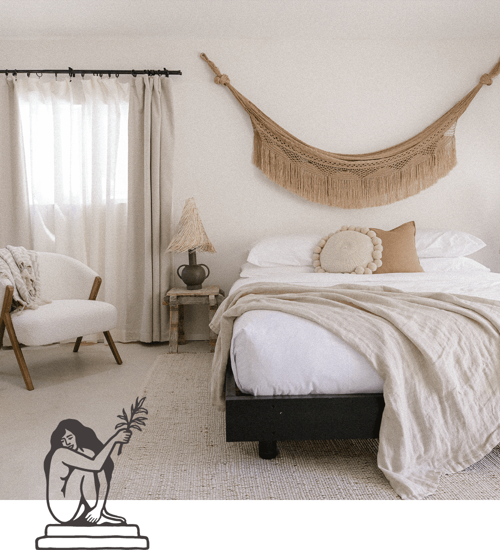 A cozy bedroom featuring a bed with neutral bedding, a decorative wall hanging, and a chair beside a wooden bedside table with a lamp.