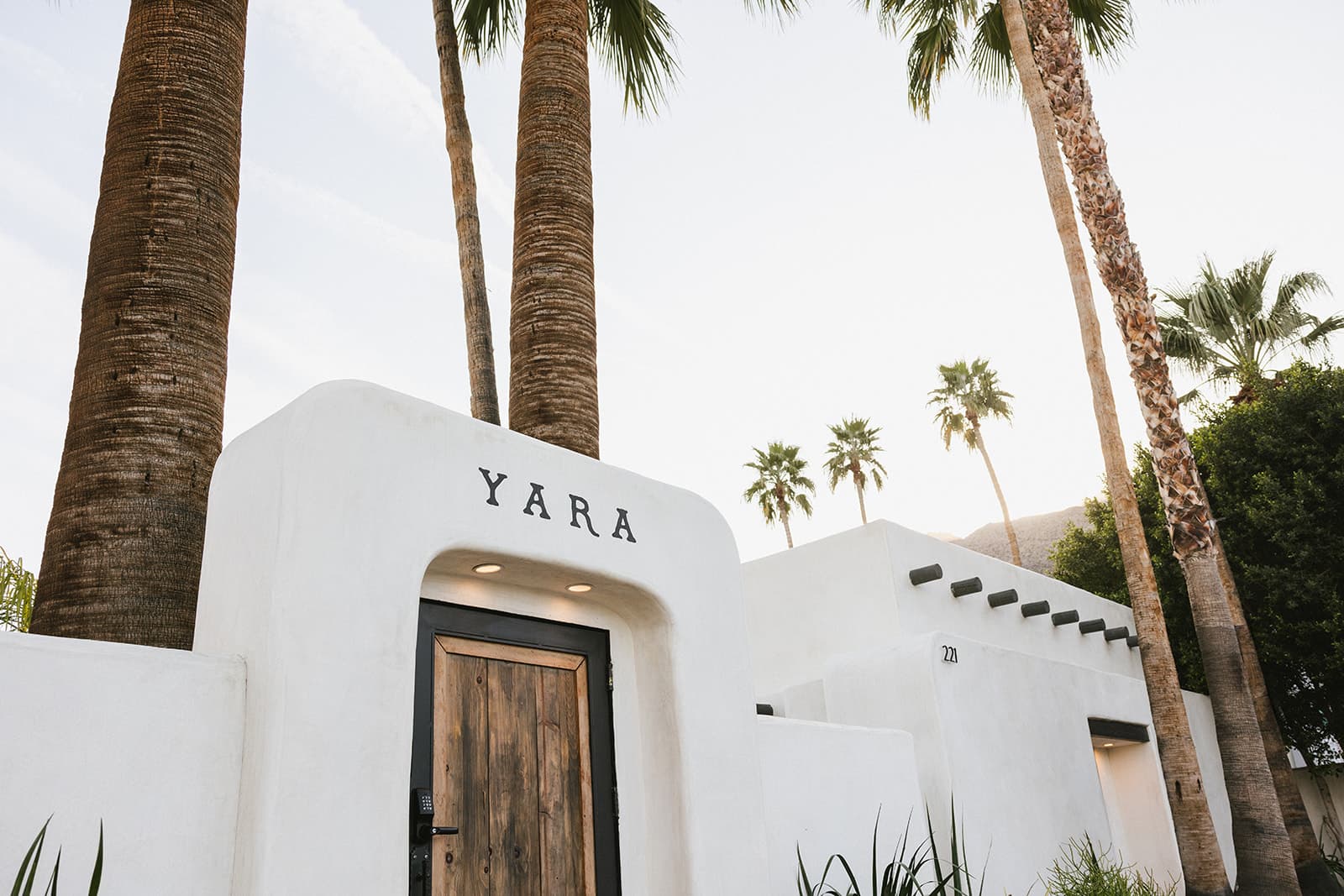 White stucco building with a wooden door and palm trees beside it, featuring the name "YARA" above the entrance. White stucco building with a wooden door and palm trees beside it, featuring the name "YARA" above the entrance.