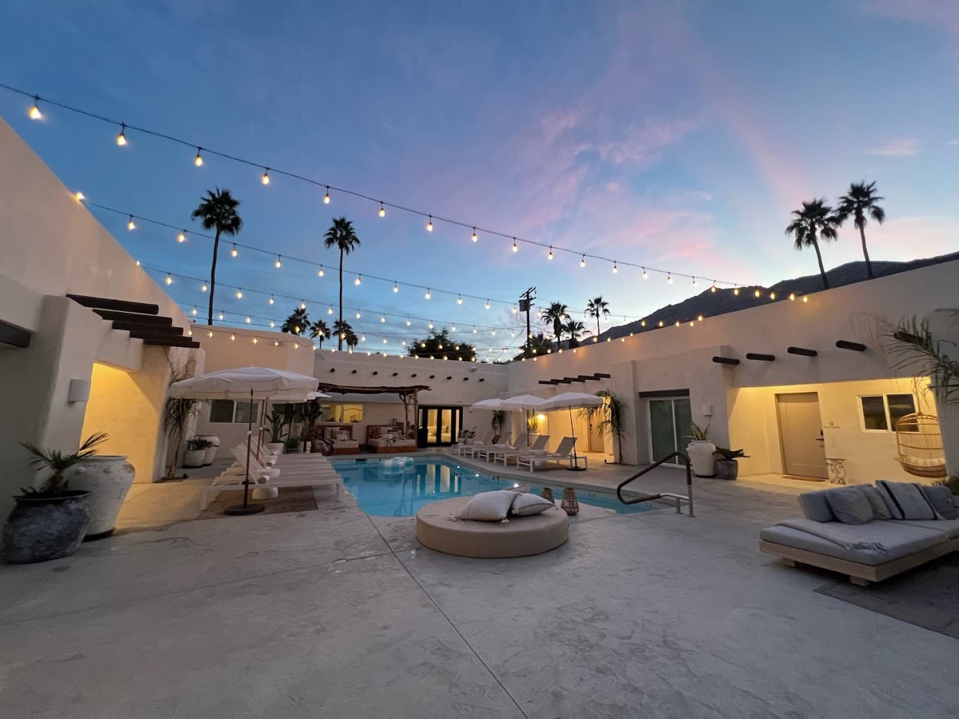 A modern patio with a pool, surrounded by palm trees and illuminated by string lights at sunset.