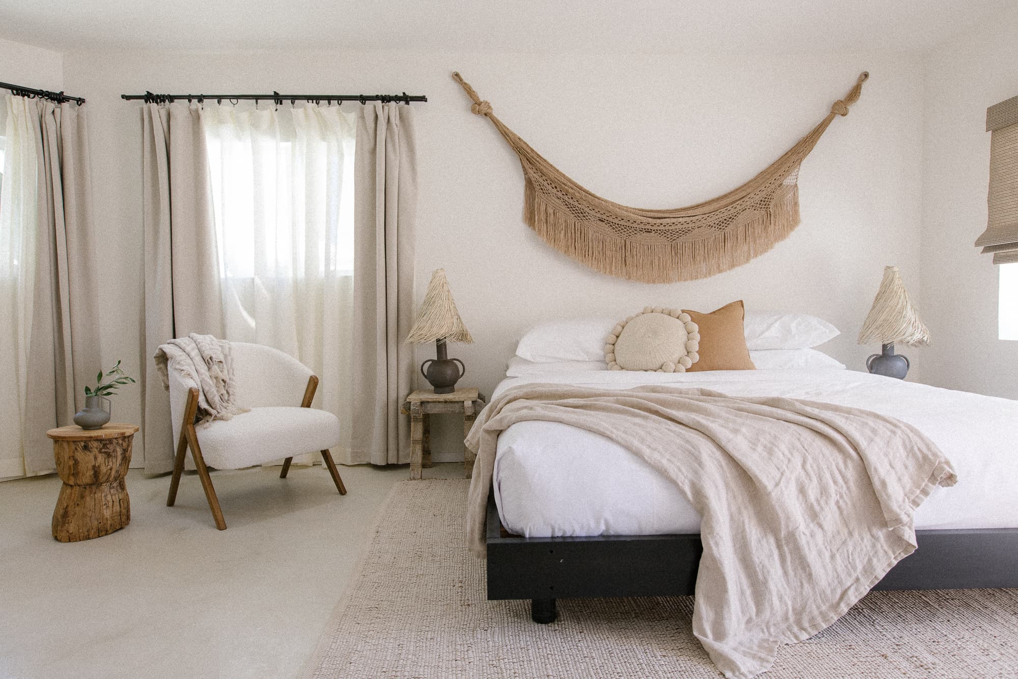 A bright, bohemian-style bedroom featuring a king bed with neutral linens and accent pillows, flanked by rustic tables and unique lamps. A woven macrame wall hanging is above the bed.