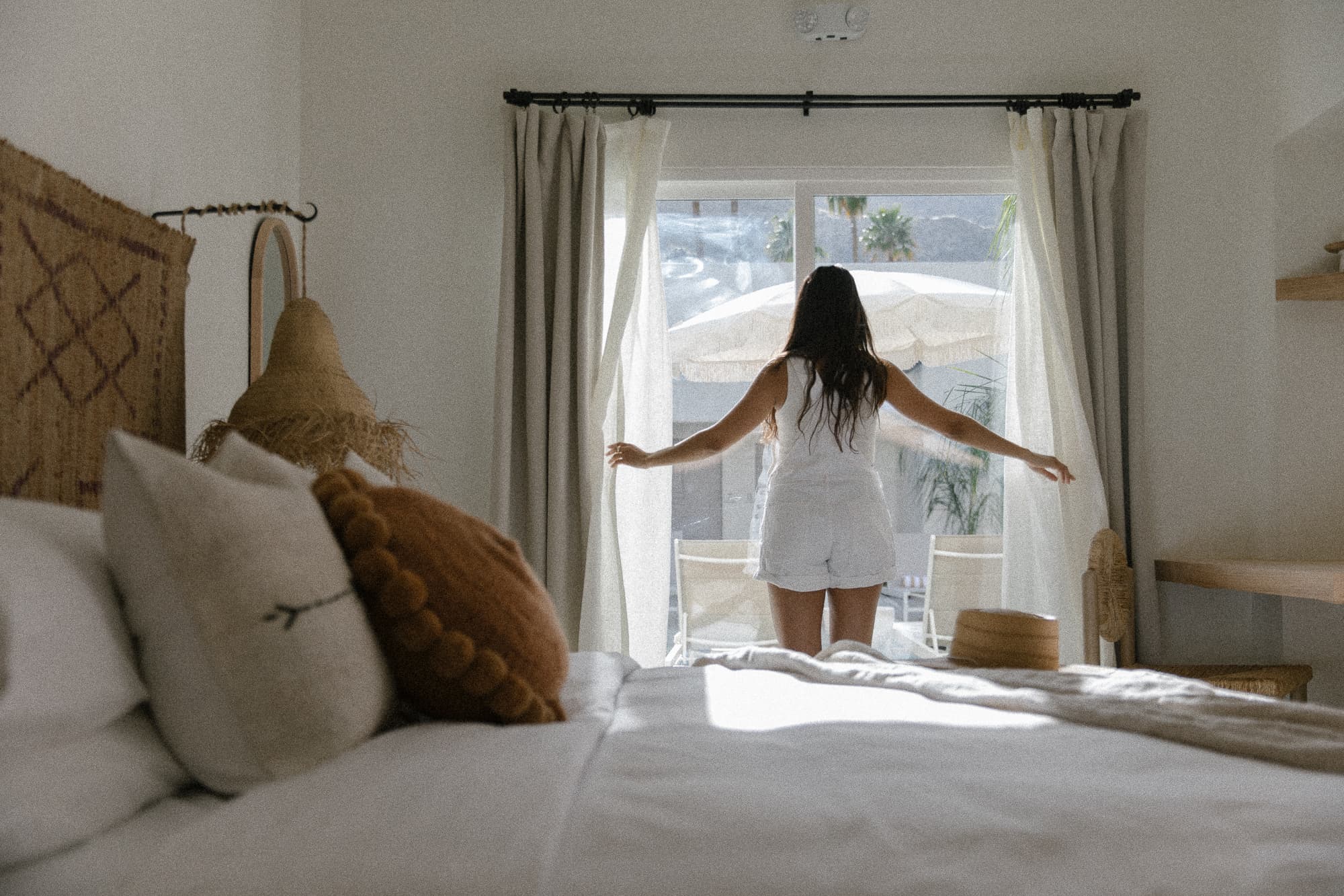 A person in a white outfit stands by a window, pulling back sheer curtains to reveal an outdoor patio.