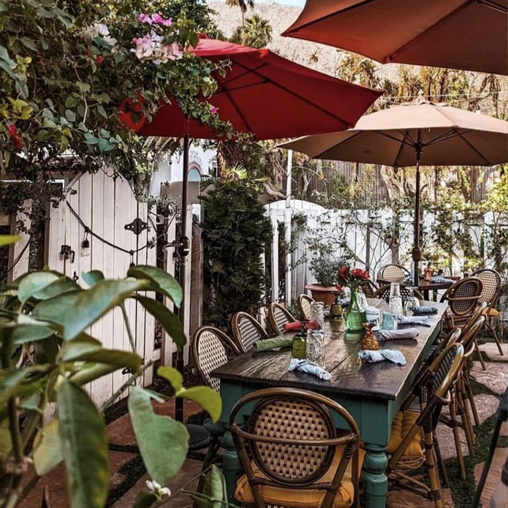 An outdoor dining area with a large table, surrounded by chairs and shaded by colorful umbrellas, amidst lush greenery.