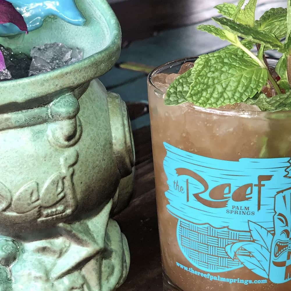 A tropical cocktail garnished with mint sits next to a decorative tiki mug filled with ice.