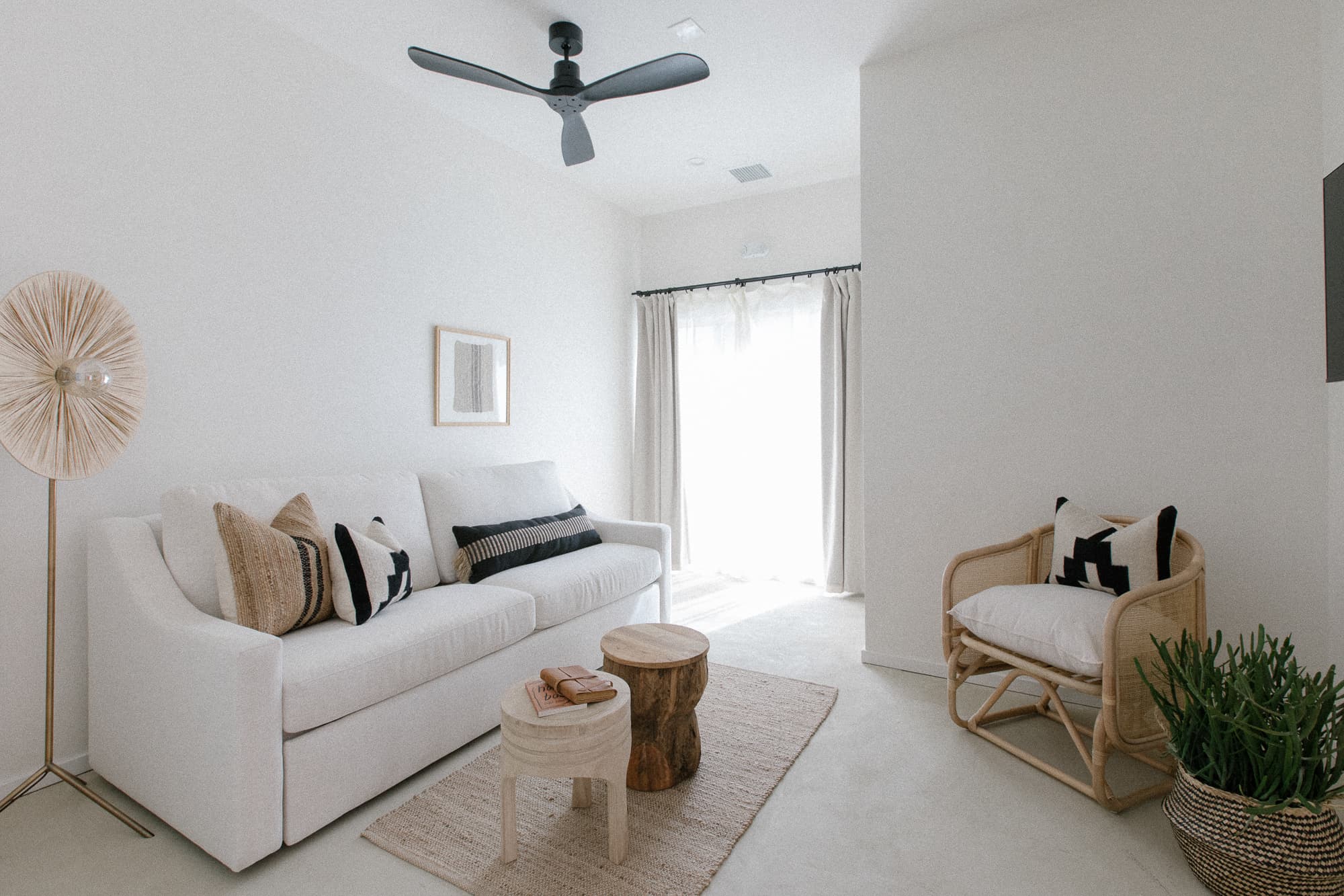 A bright, minimalist living space featuring a white sofa with neutral pillows, natural wood tables, a rattan chair, a woven floor lamp, and a black ceiling fan.