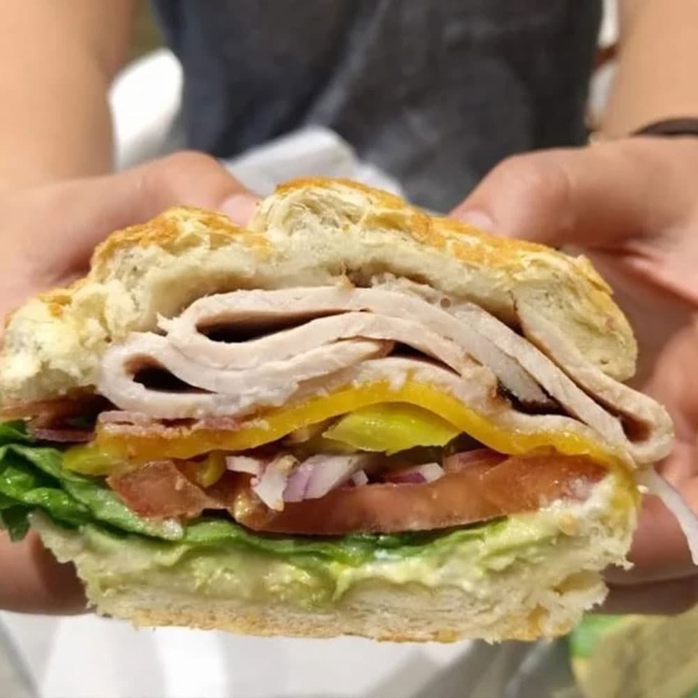 A person holds a sandwich filled with turkey, cheese, lettuce, tomato, onions, and pickles, showcasing its layers.