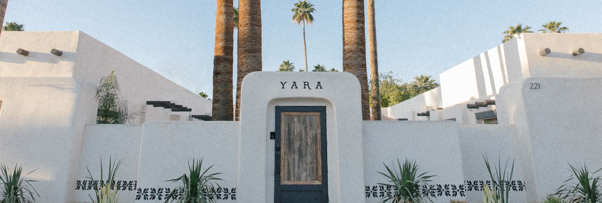 Entrance to a whitewashed building named "Yara," framed by tall palm trees and greenery.