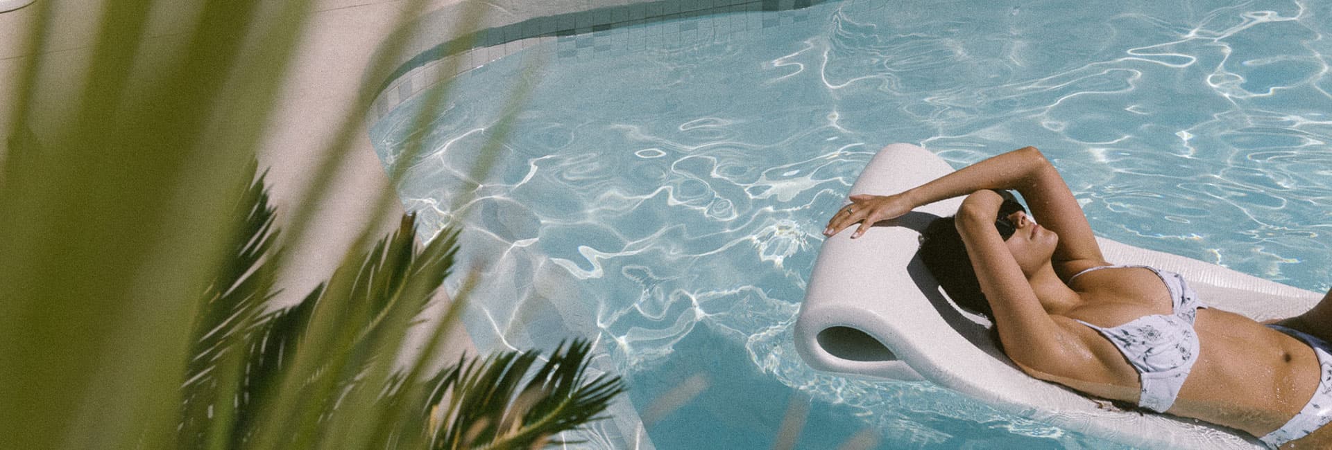 A person relaxes on a pool float in a sunlit swimming pool surrounded by greenery.