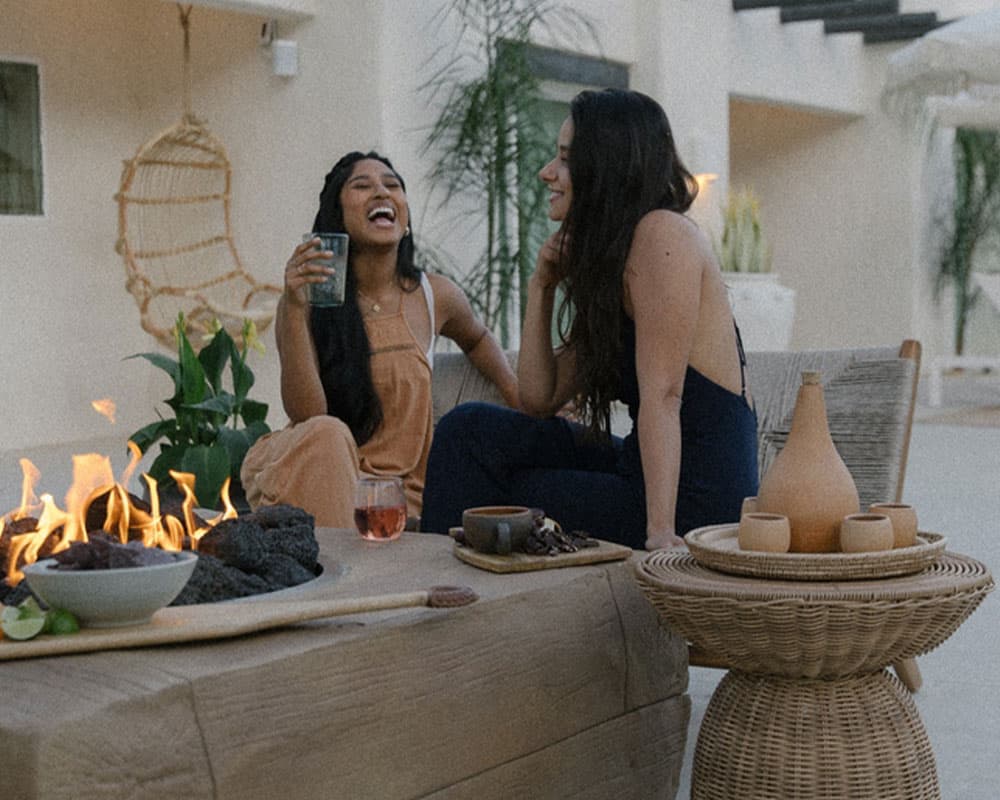 Two women laugh and enjoy drinks around a cozy fire pit in a stylish outdoor setting.